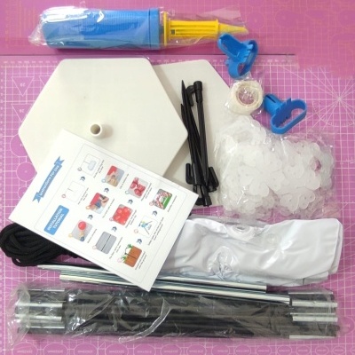 Kit de montagem com peças variadas como base branca, hastes, suportes azuis, bomba de ar e manual sobre superfície rosa