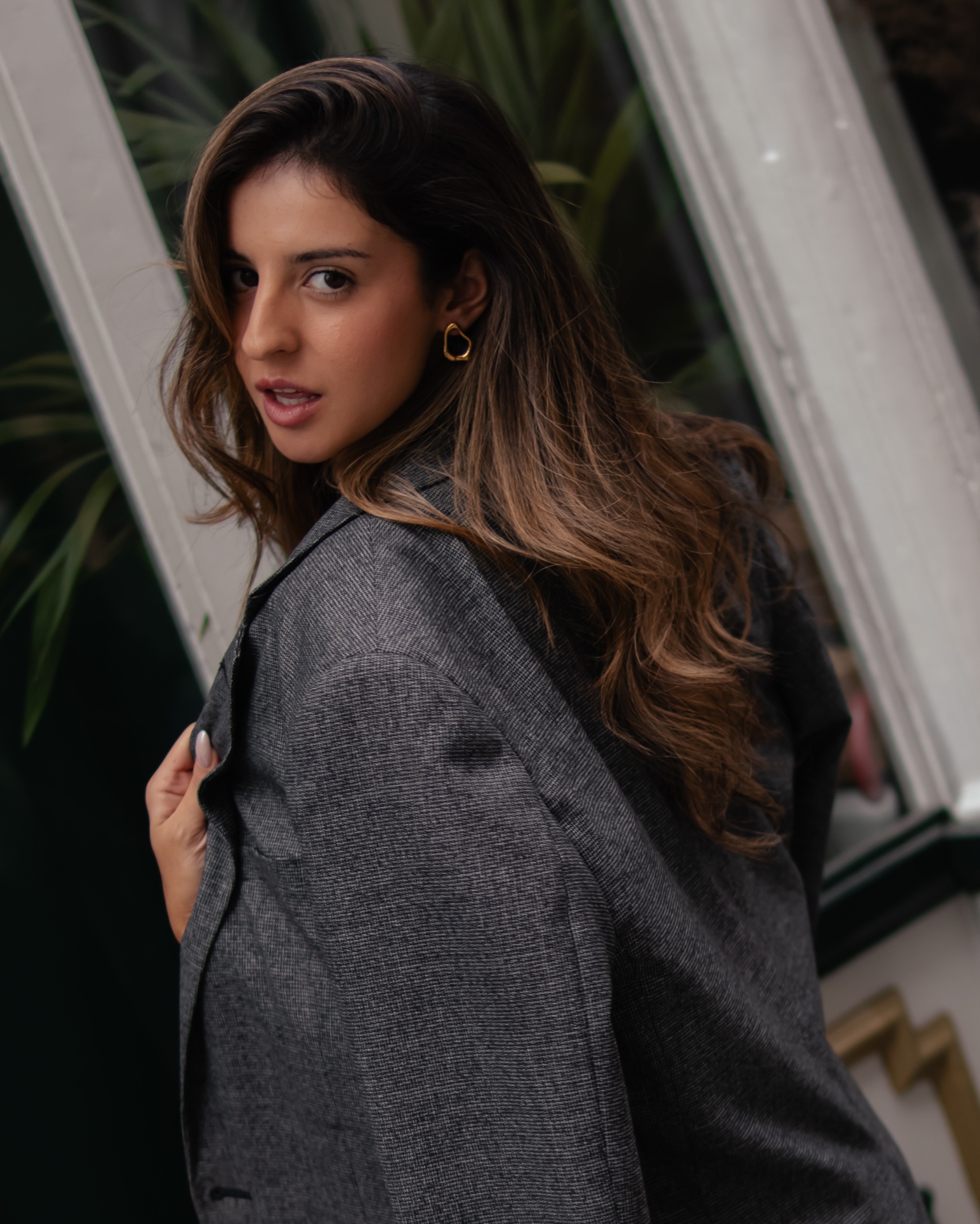 Mulher de casaco cinza escuro em frente a espelho com plantas ao fundo