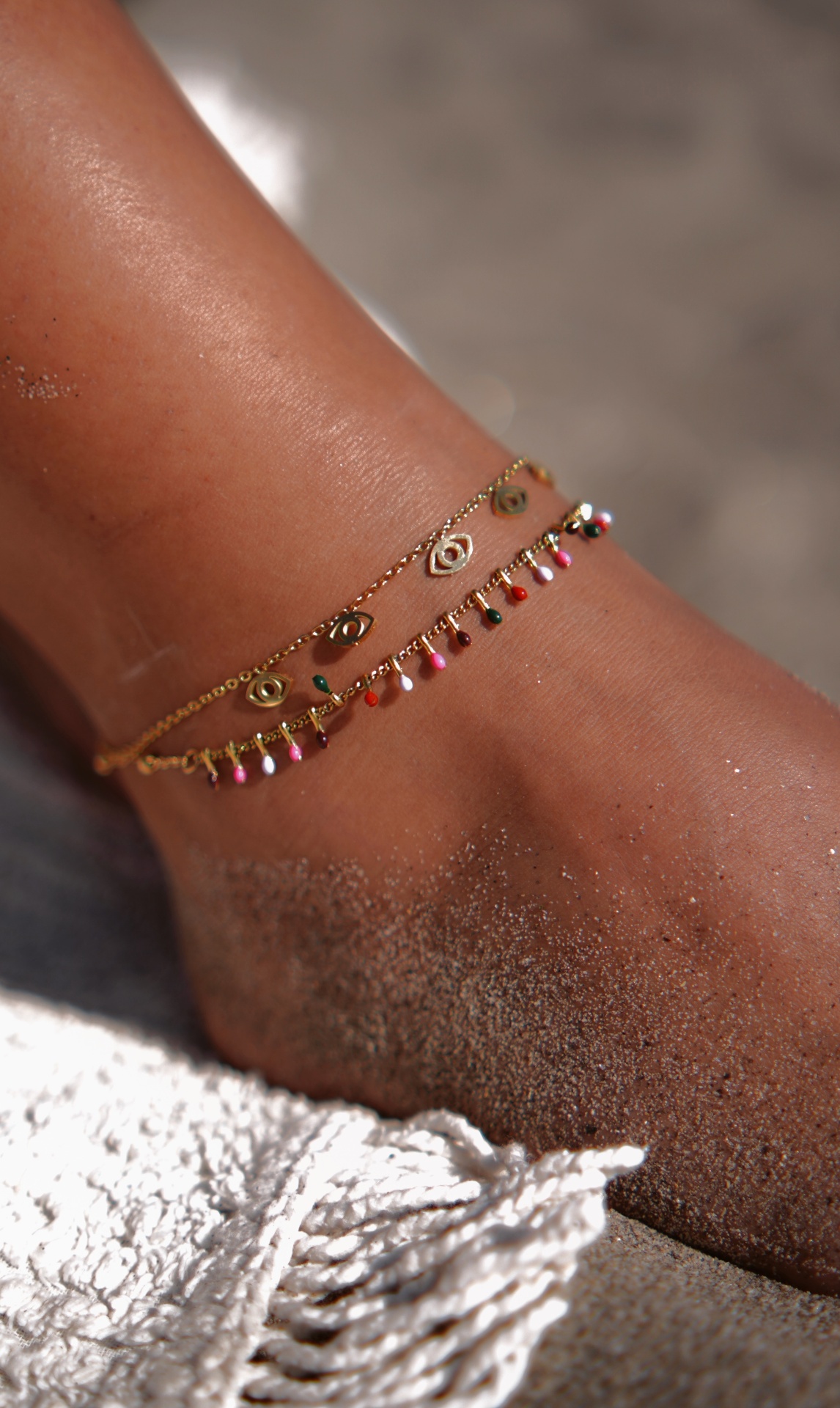 Duas tornozeleiras douradas delicadas com pendentes coloridos e amuletos em tornozelo arenoso na praia