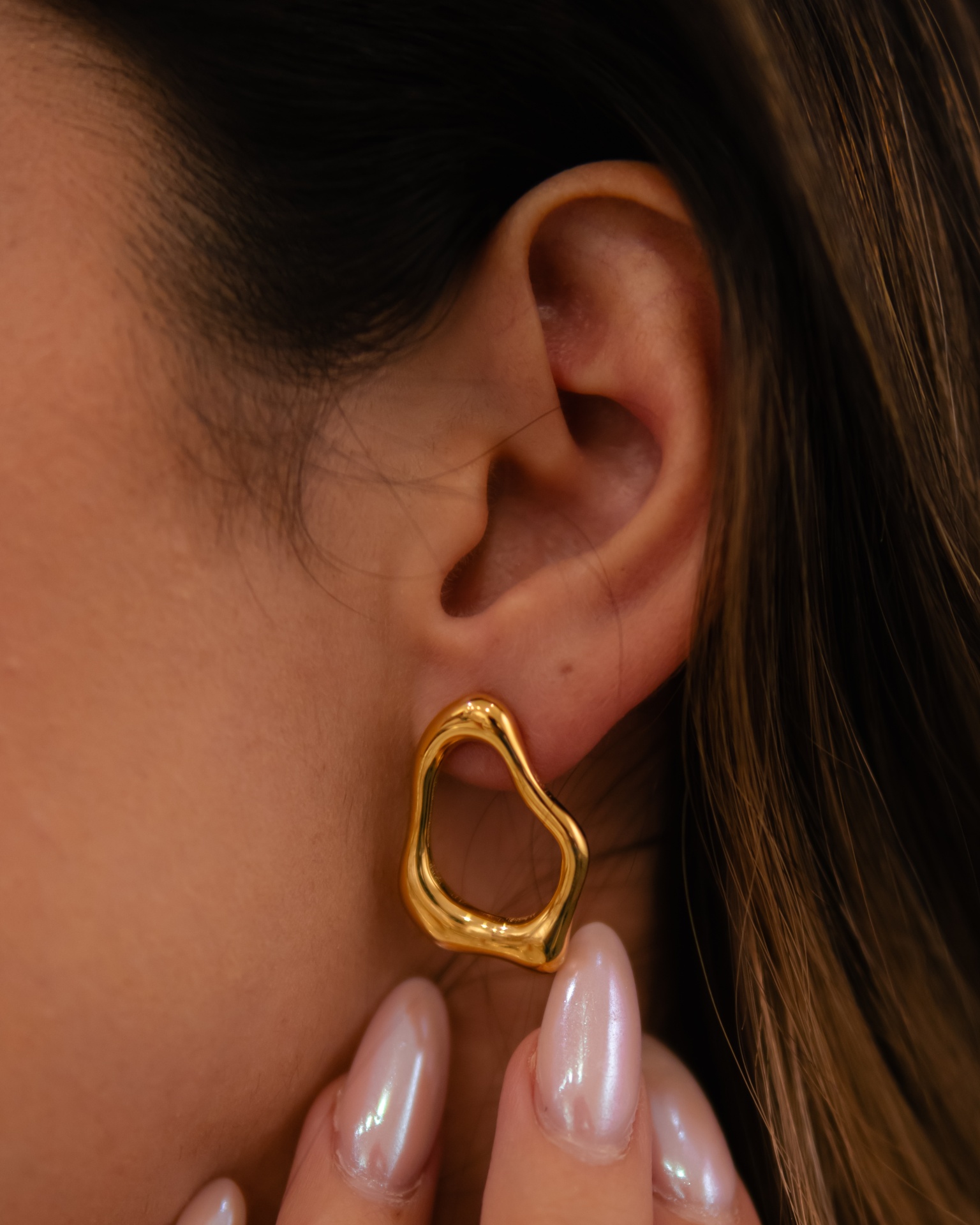 Brinco de ouro irregular usado na orelha de uma mulher com unhas peroladas.