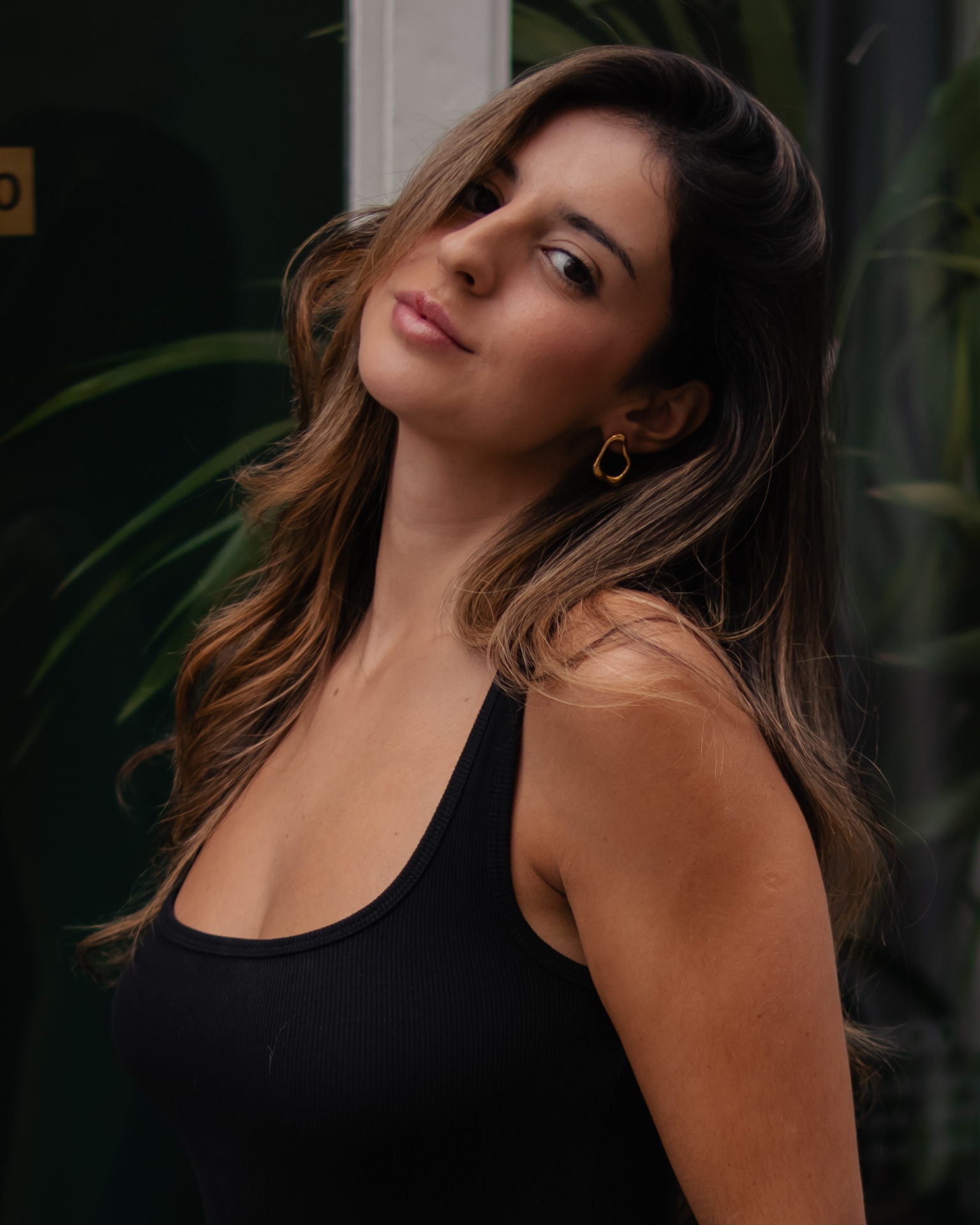 Mulher com top preto e brincos dourados em fundo verde com plantas.