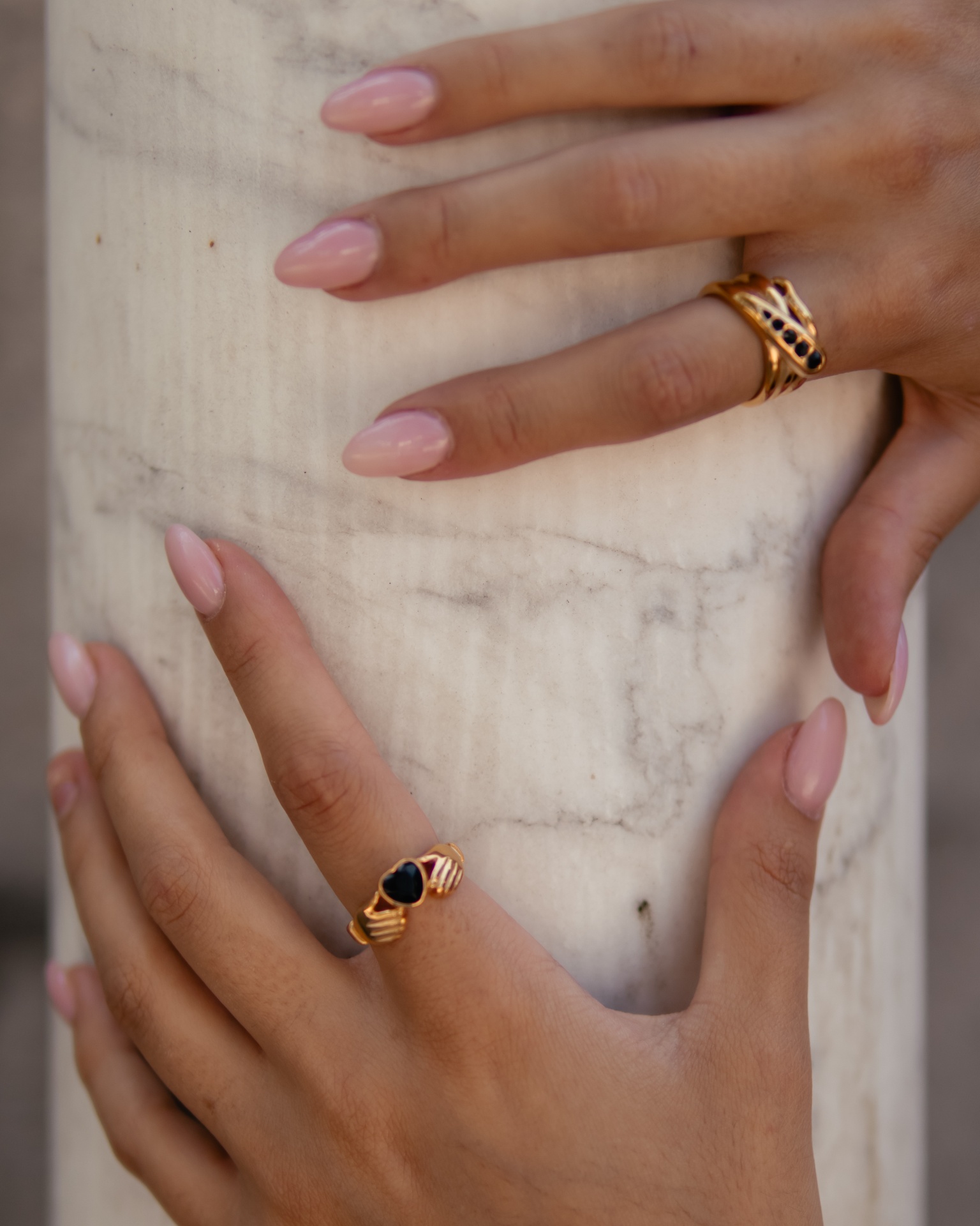 Mãos femininas com anéis dourados e unhas cor-de-rosa segurando coluna de mármore branca