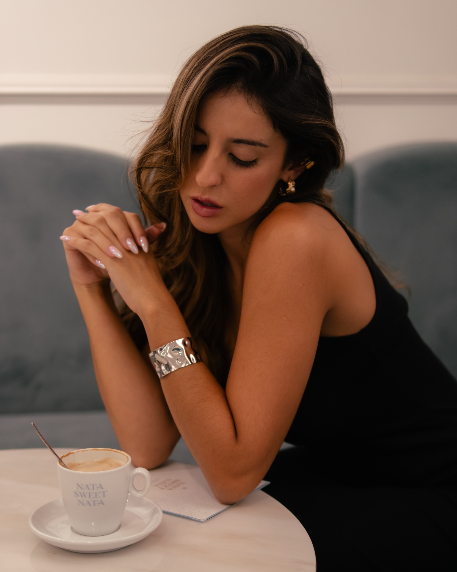 Mulher com vestido preto e pulseira prateada junto a uma chávena de café branca numa mesa branca