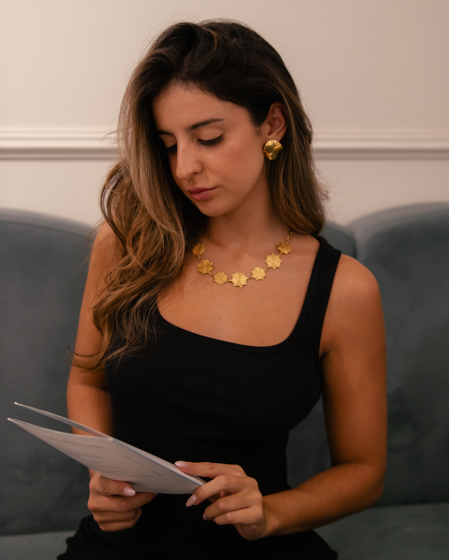 Mulher de vestido preto com colar e brincos dourados, sentada e lendo papel