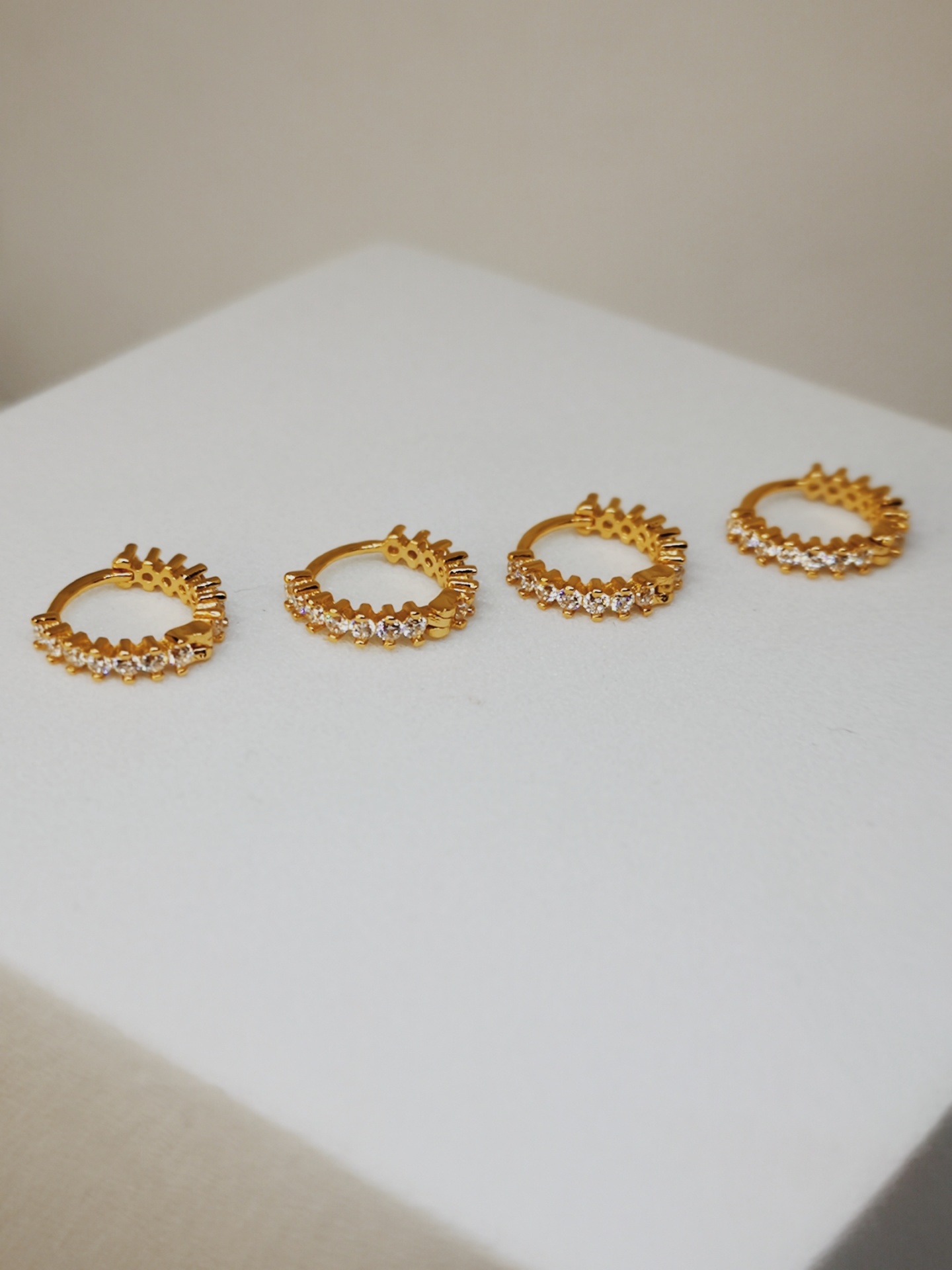 Quatro argolas douradas com pedras claras e detalhes em forma de picos sobre superfície branca