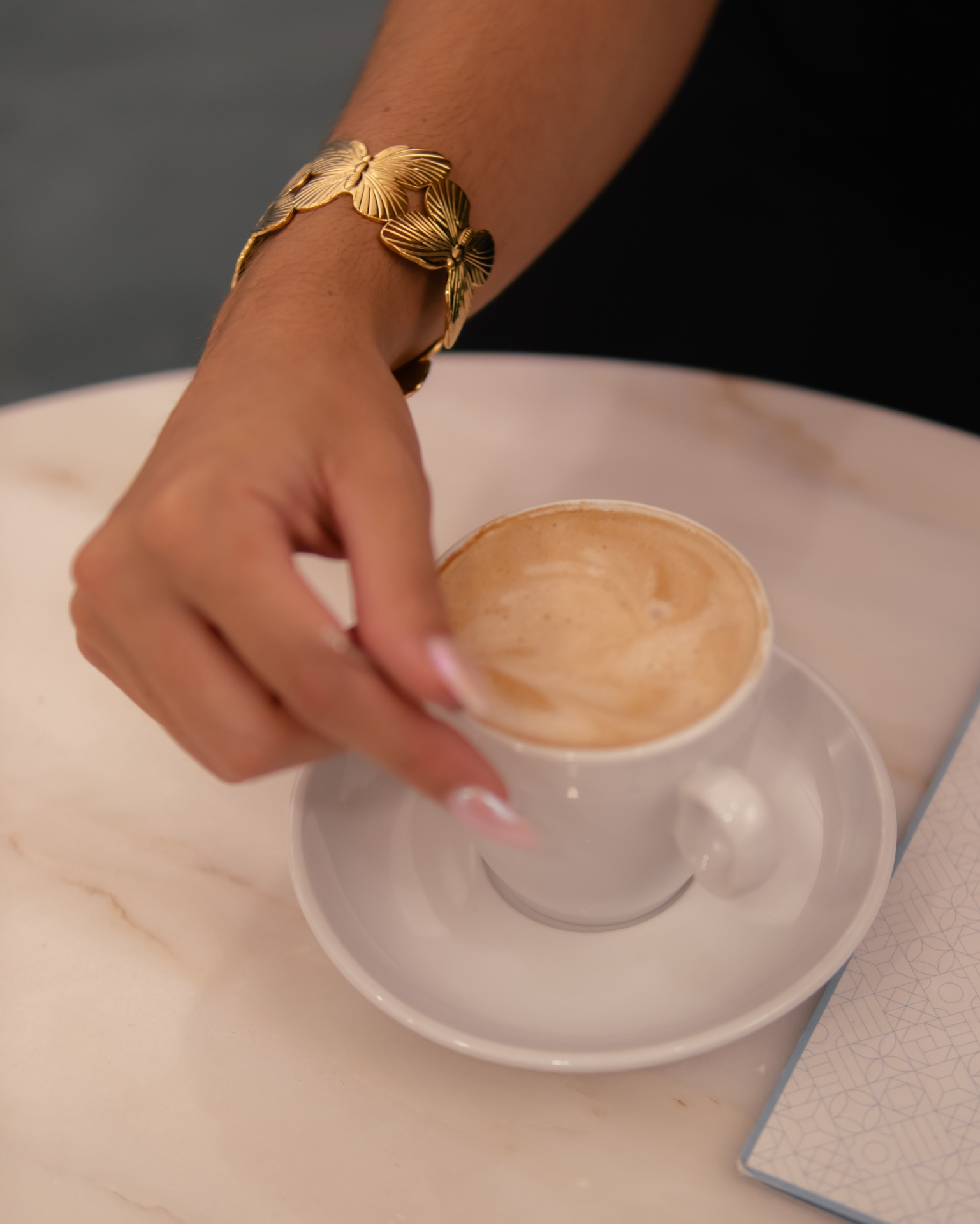 Braço com pulseira dourada segurando chávena branca de café em mesa de mármore