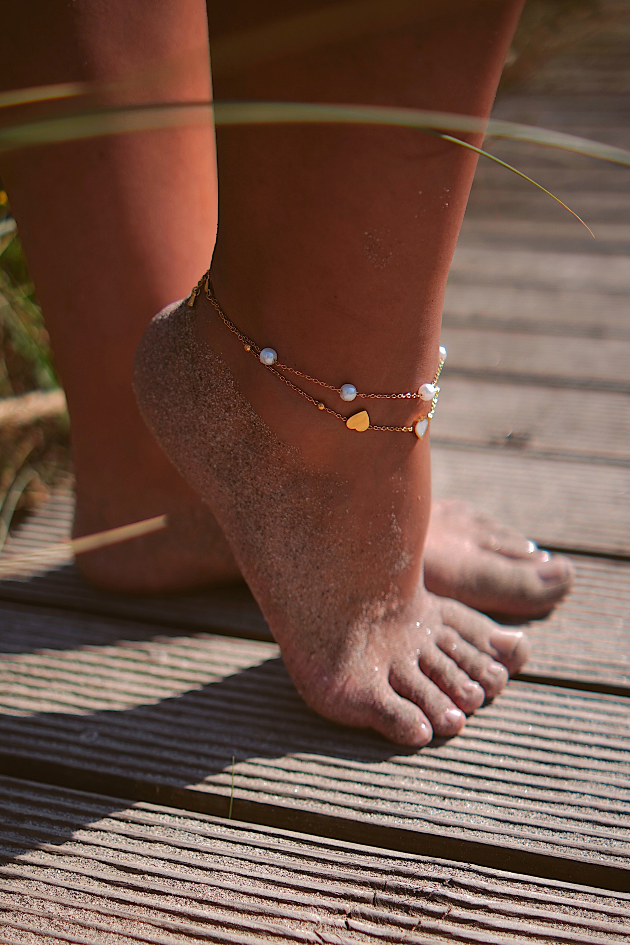 Pé com tornozeleiras douradas com pérolas e coração em passadiço de madeira