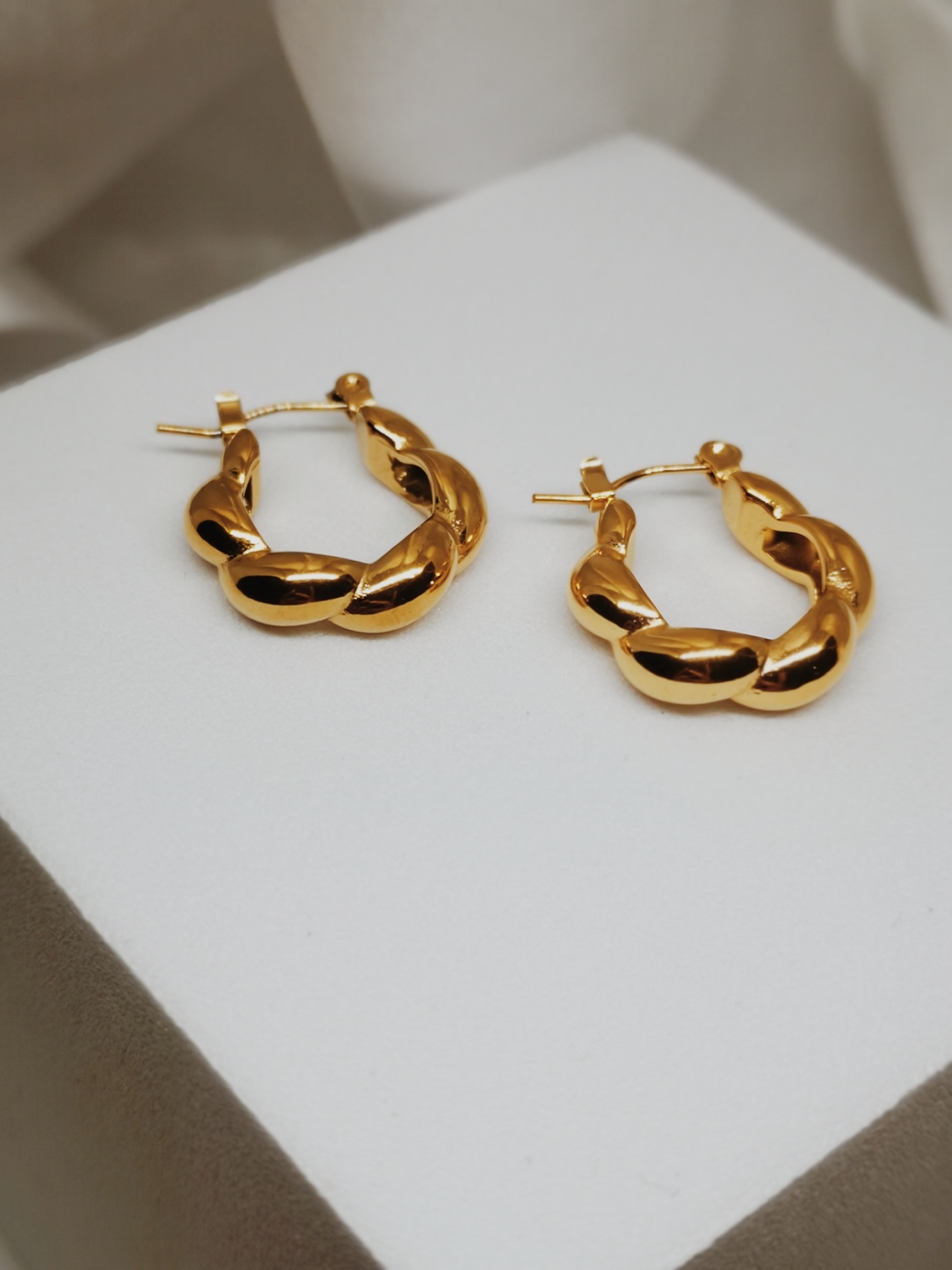 Brincos de argola dourados com design trançado sobre fundo branco e bege