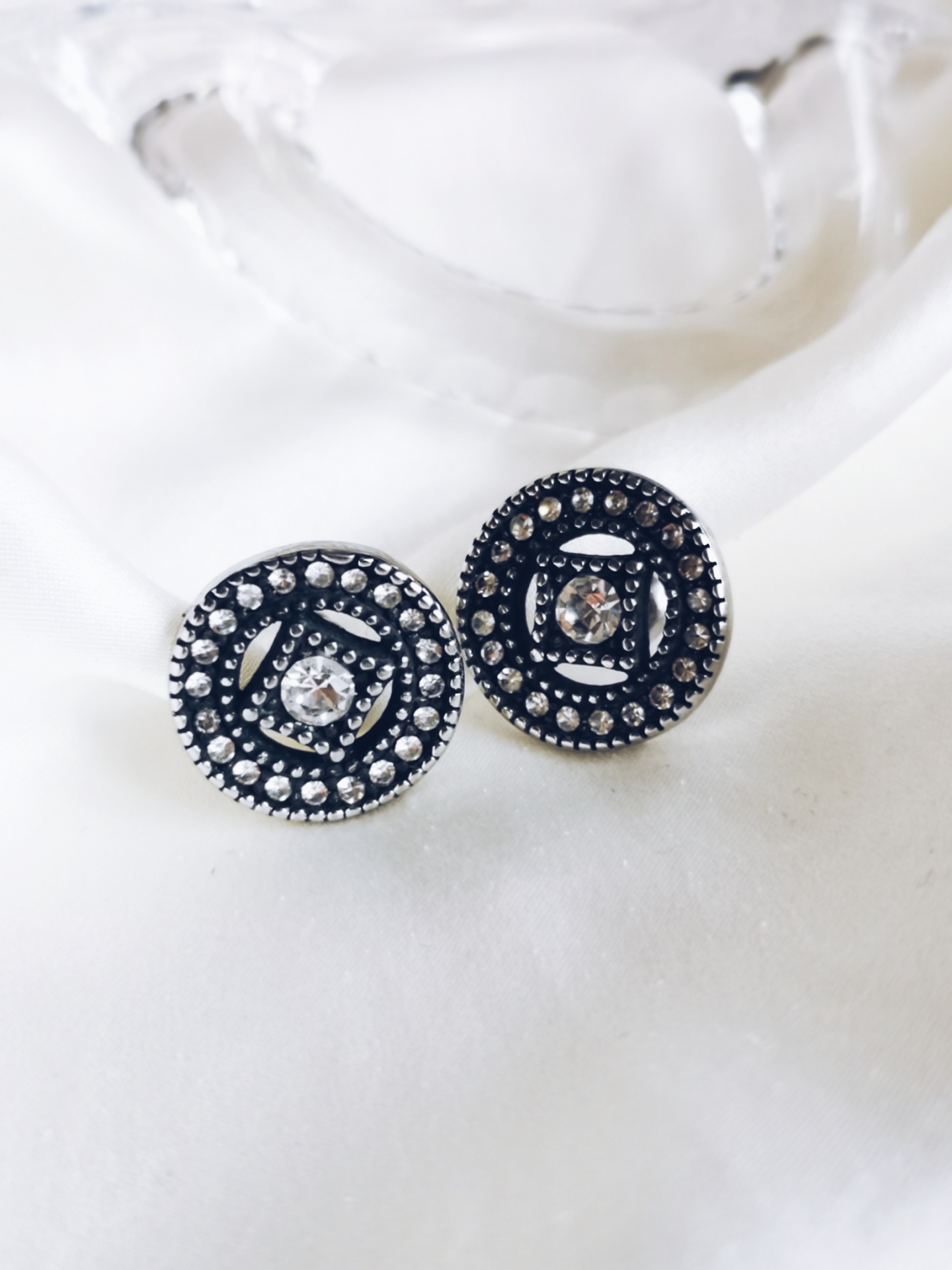 Brincos redondos pretos com detalhes prateados e pedra central sobre tecido branco