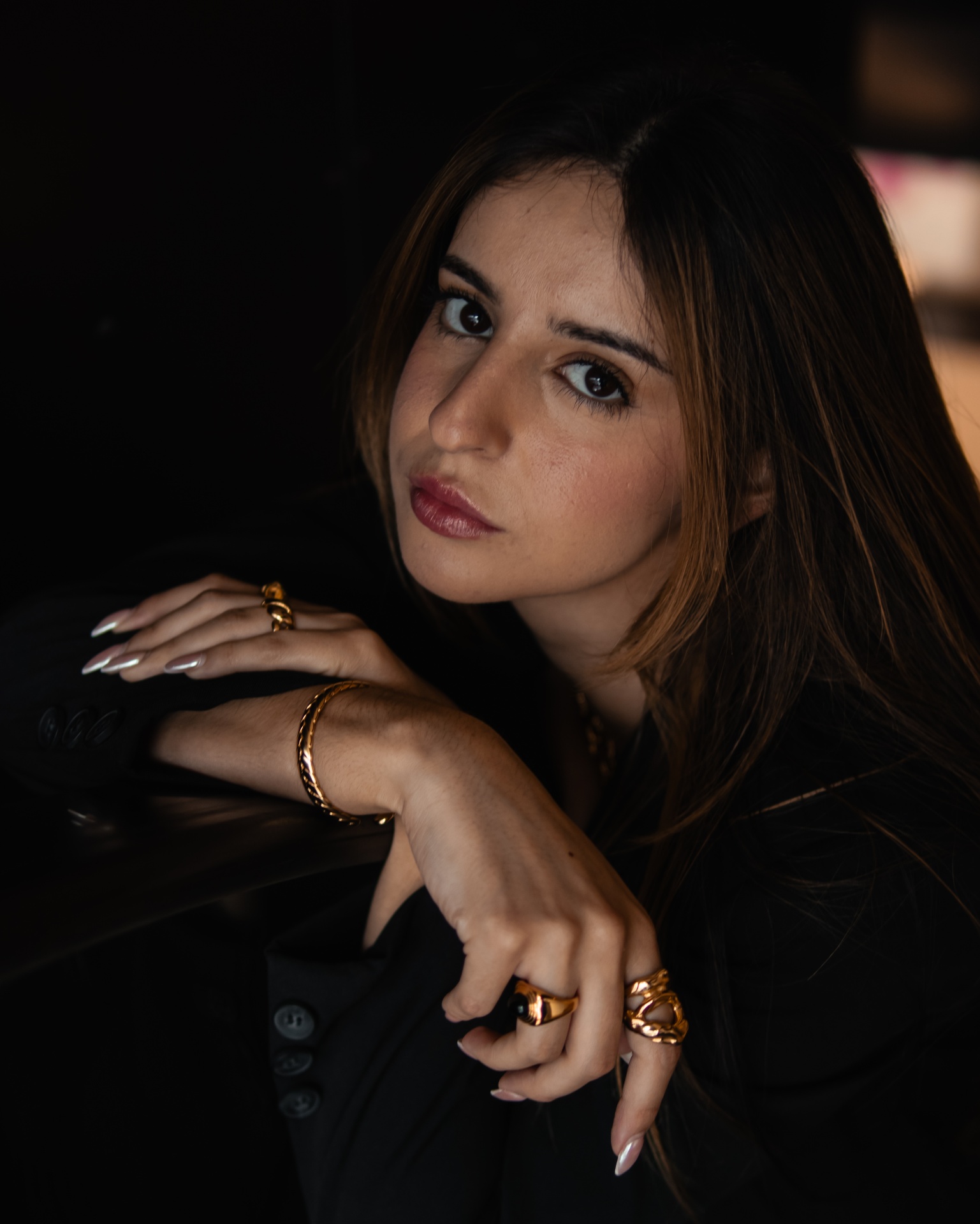 Mulher com acessórios dourados e casaco preto em fundo escuro