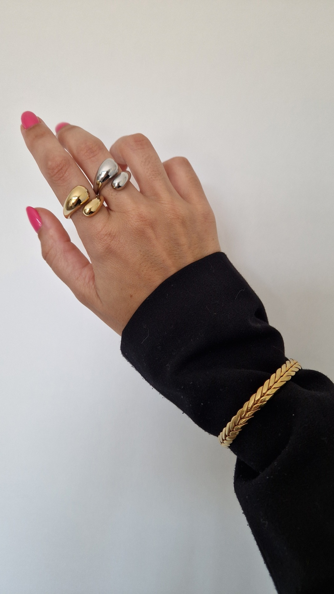 Mão feminina com três anéis metálicos e pulseira dourada, unhas rosas e manga preta