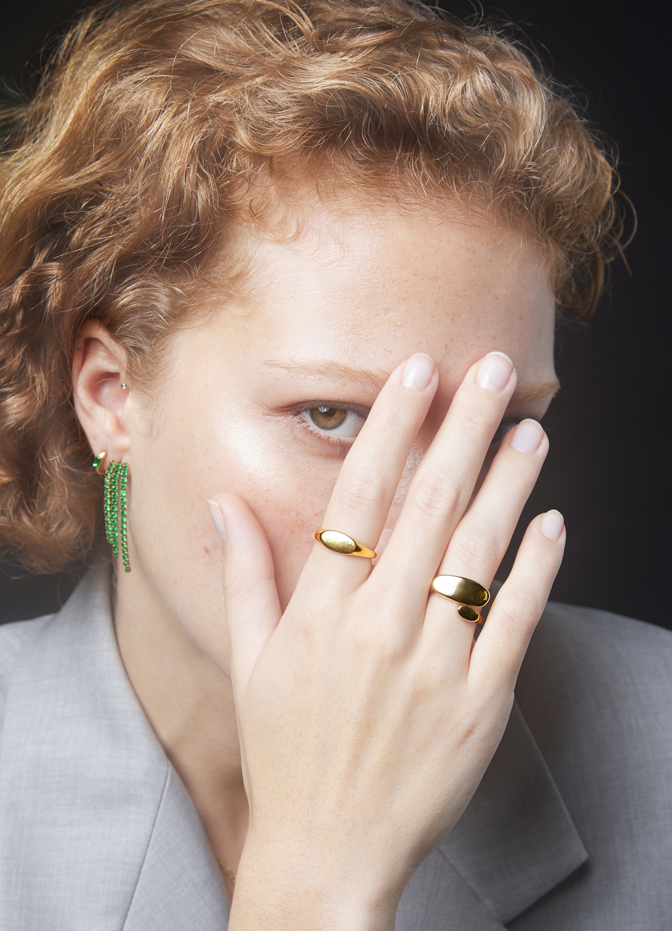 Mulher com brincos pendentes verdes e anéis dourados cobre parcialmente o rosto com a mão esquerda
