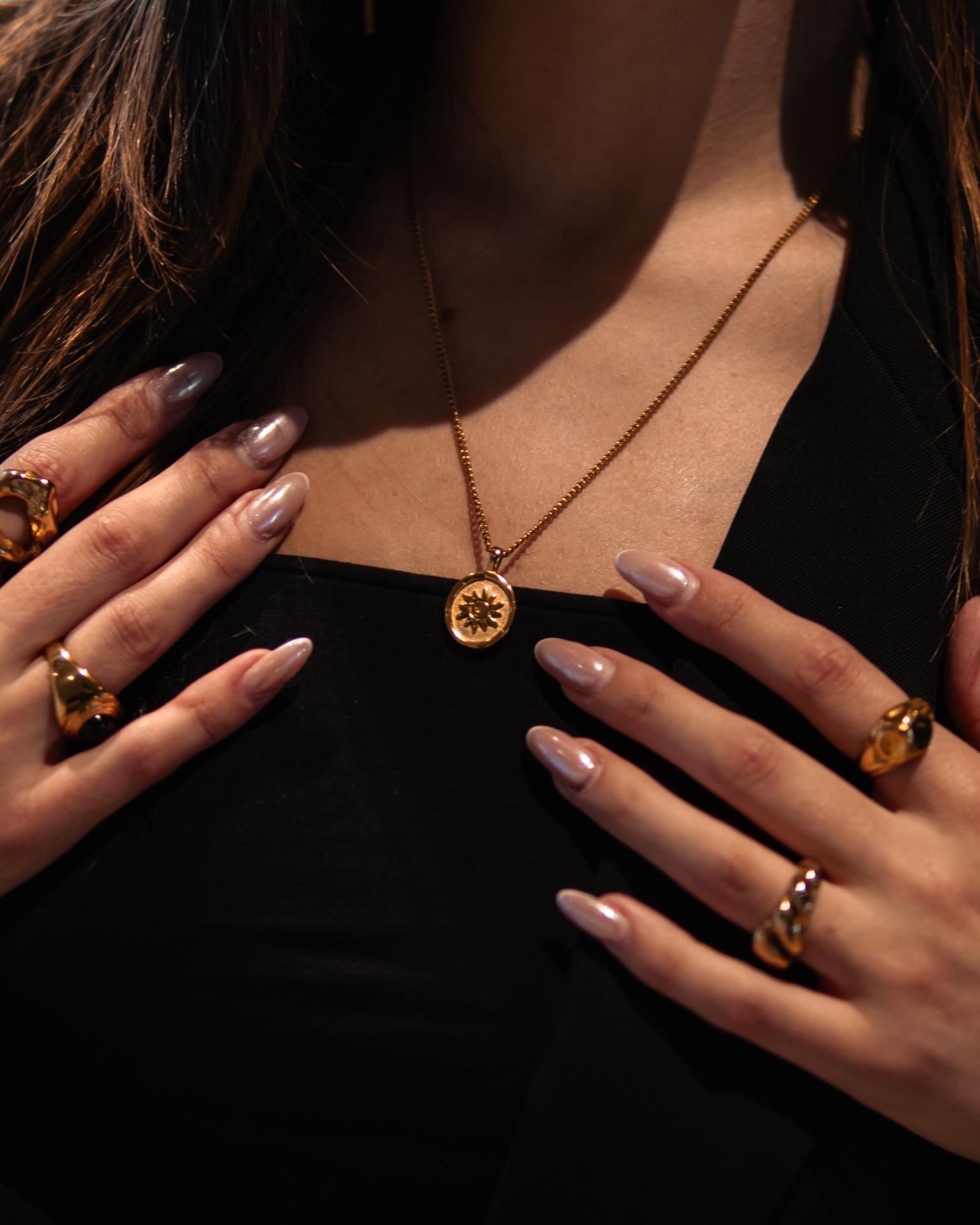 Colar dourado com medalhão redondo e anéis dourados com pedras castanhas numa pessoa com roupa preta