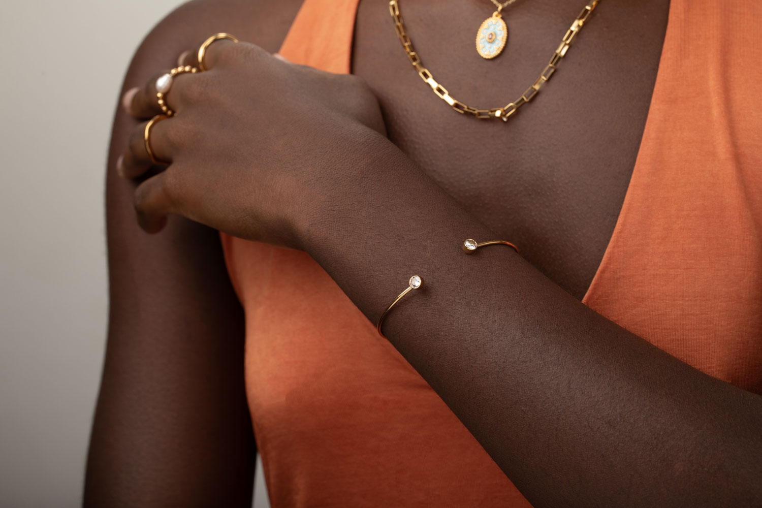 Pulseira aberta dourada com pedras e anéis dourados usados por pessoa com pele escura e t-shirt laranja.