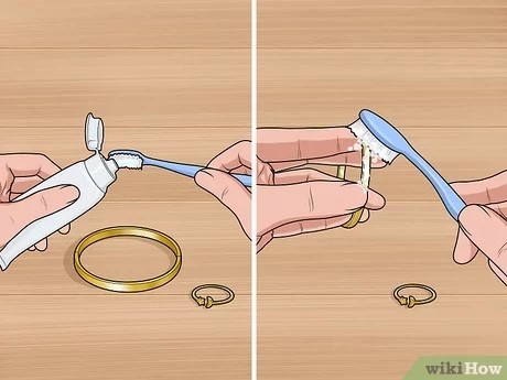 Mãos a escovar uma joia dourada com uma escova de dentes azul e pasta de dentes numa superfície de madeira.