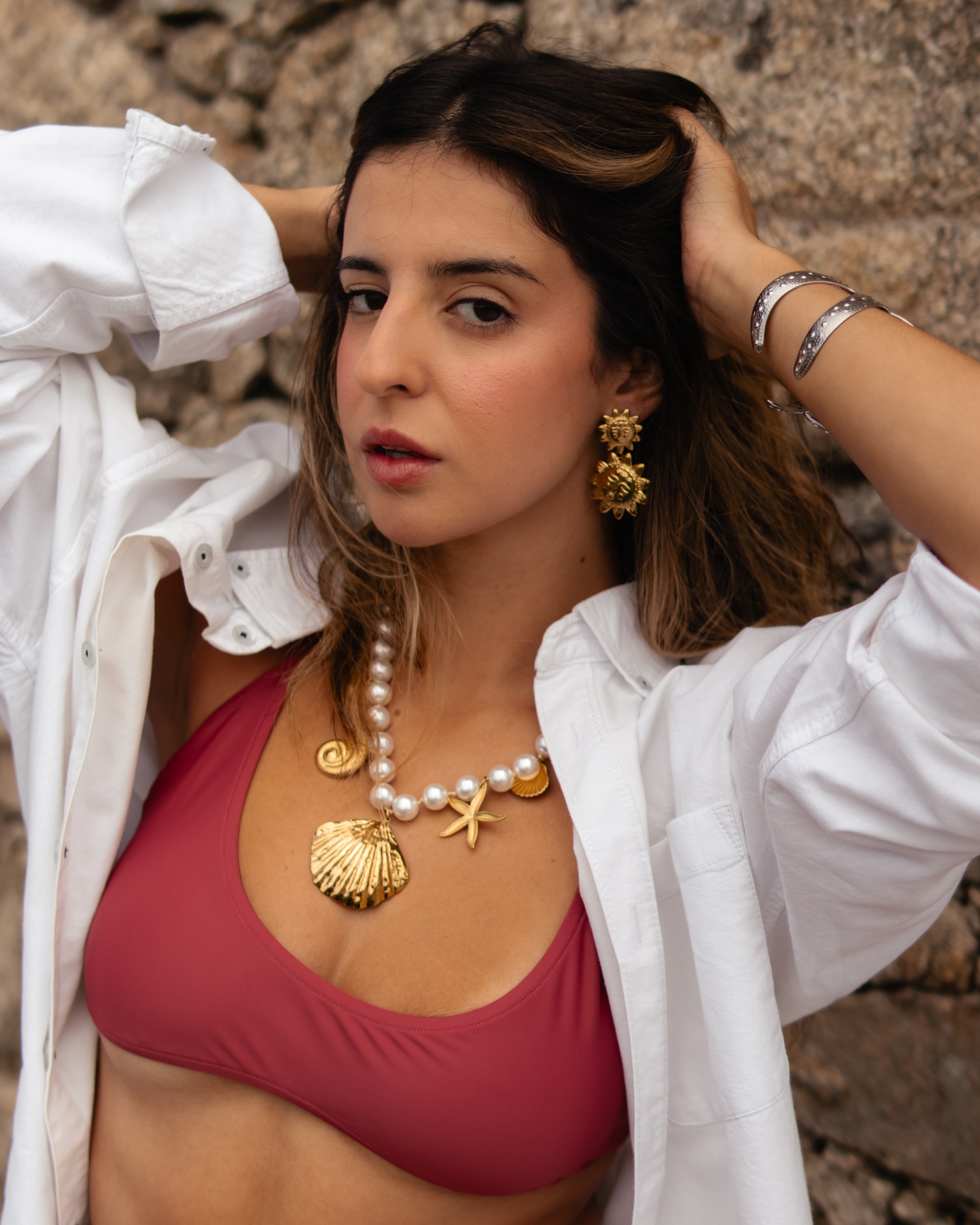 Mulher com colar de pérolas e pendente dourado, brincos dourados, top vinho e camisa branca