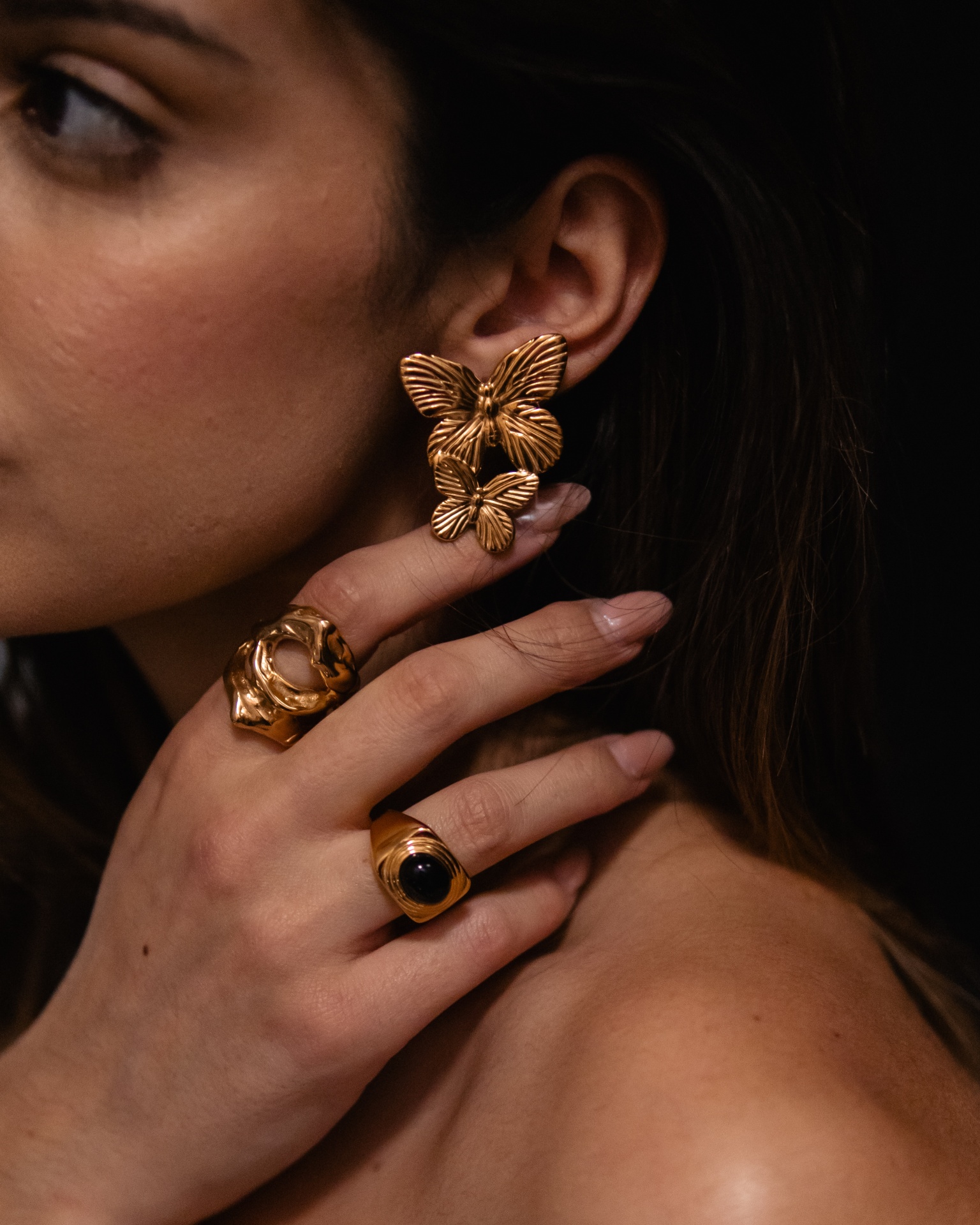 Brincos dourados de borboleta e anéis dourados numa mão junto ao rosto de mulher.