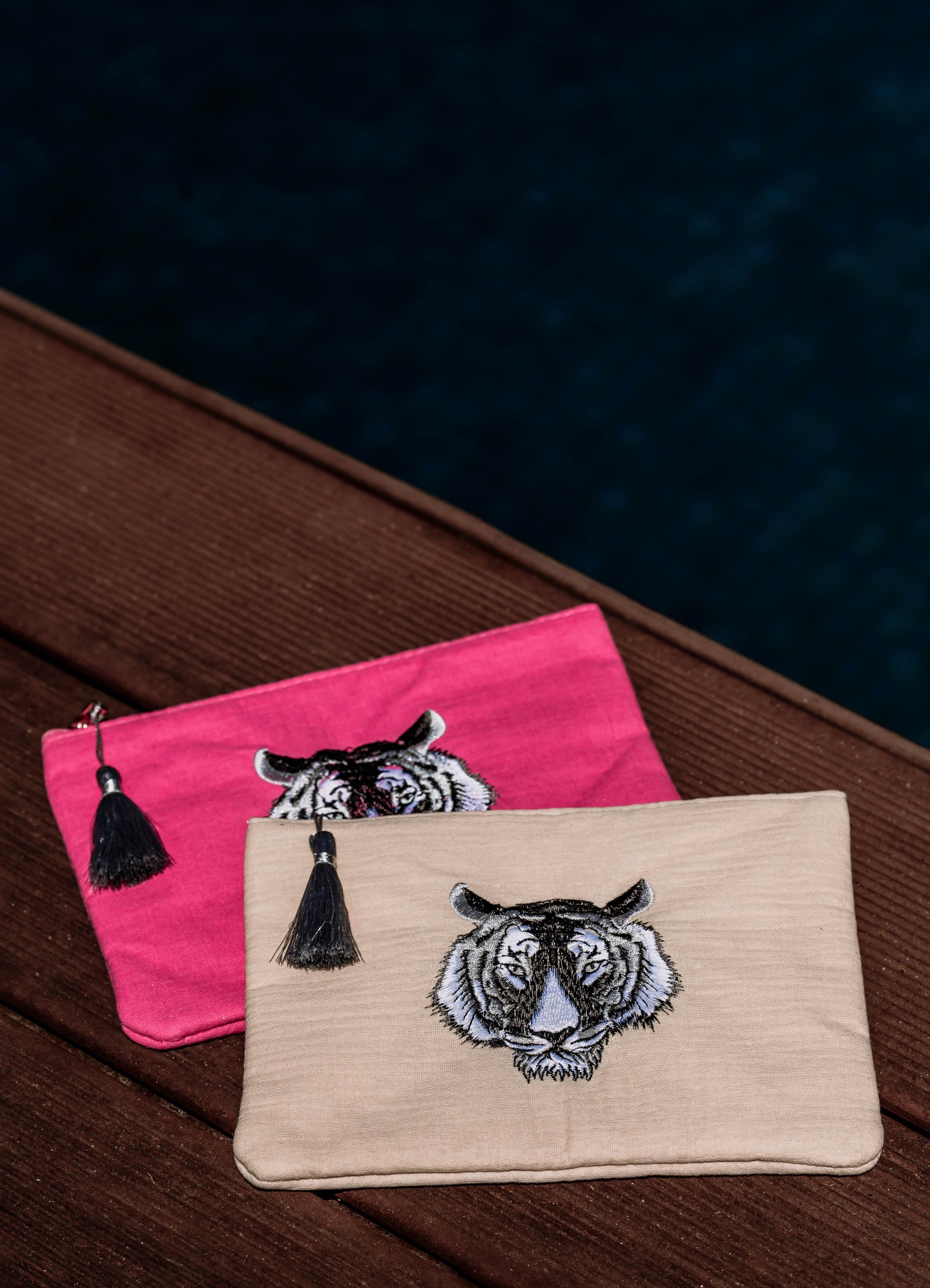 Duas bolsas coloridas com bordado de tigre e tassel preto sobre madeira ao lado de piscina