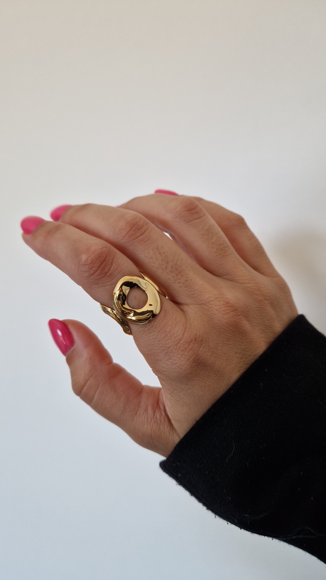 Anel dourado irregular e oco no dedo de mão com unhas rosas e manga preta