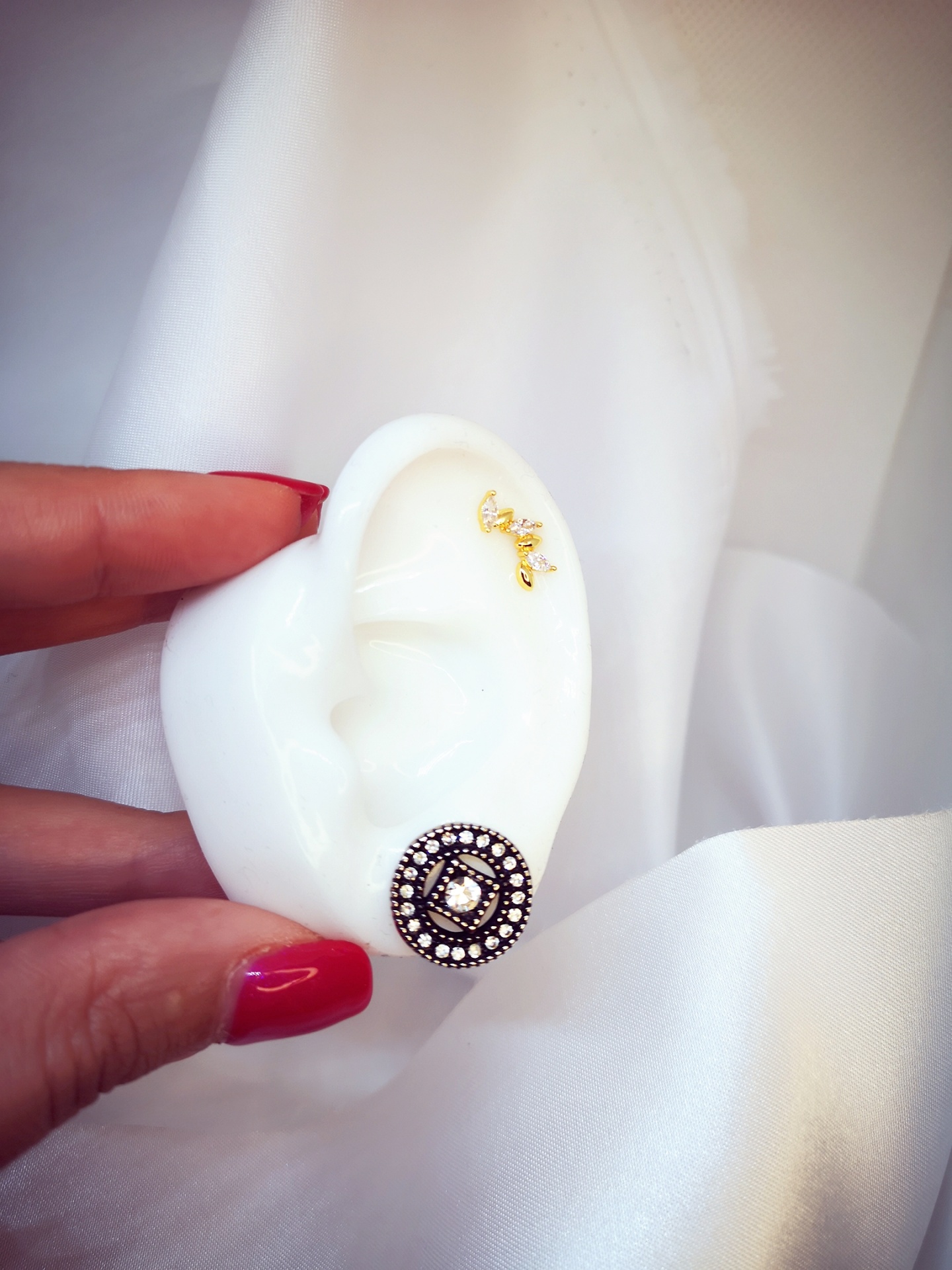 Modelo de orelha com dois brincos, um dourado pequeno e outro redondo com pedras, segurado por mão com unhas vermelhas