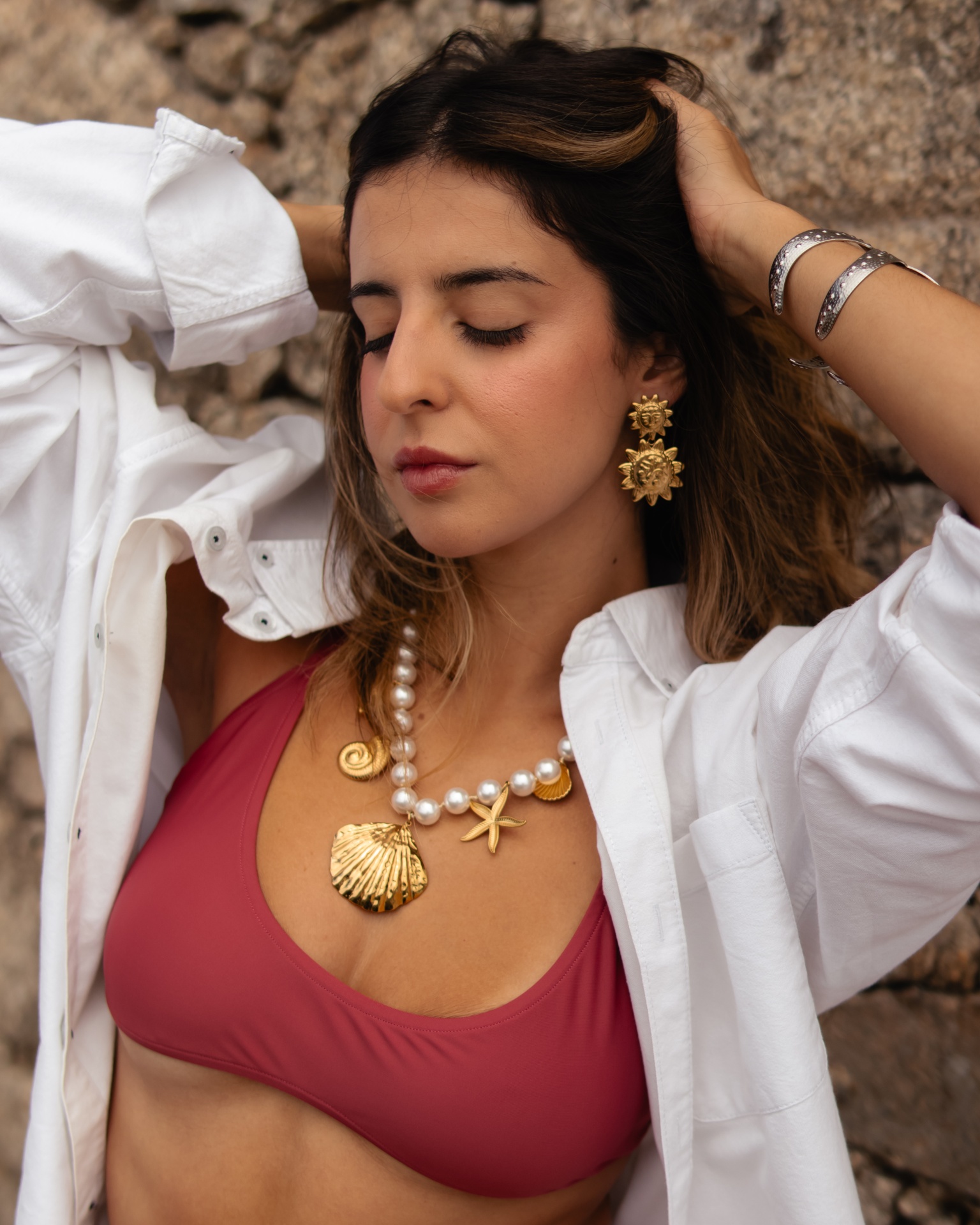 Mulher com colar e brincos dourados, pulseiras, top vermelho e camisa branca em fundo de pedra