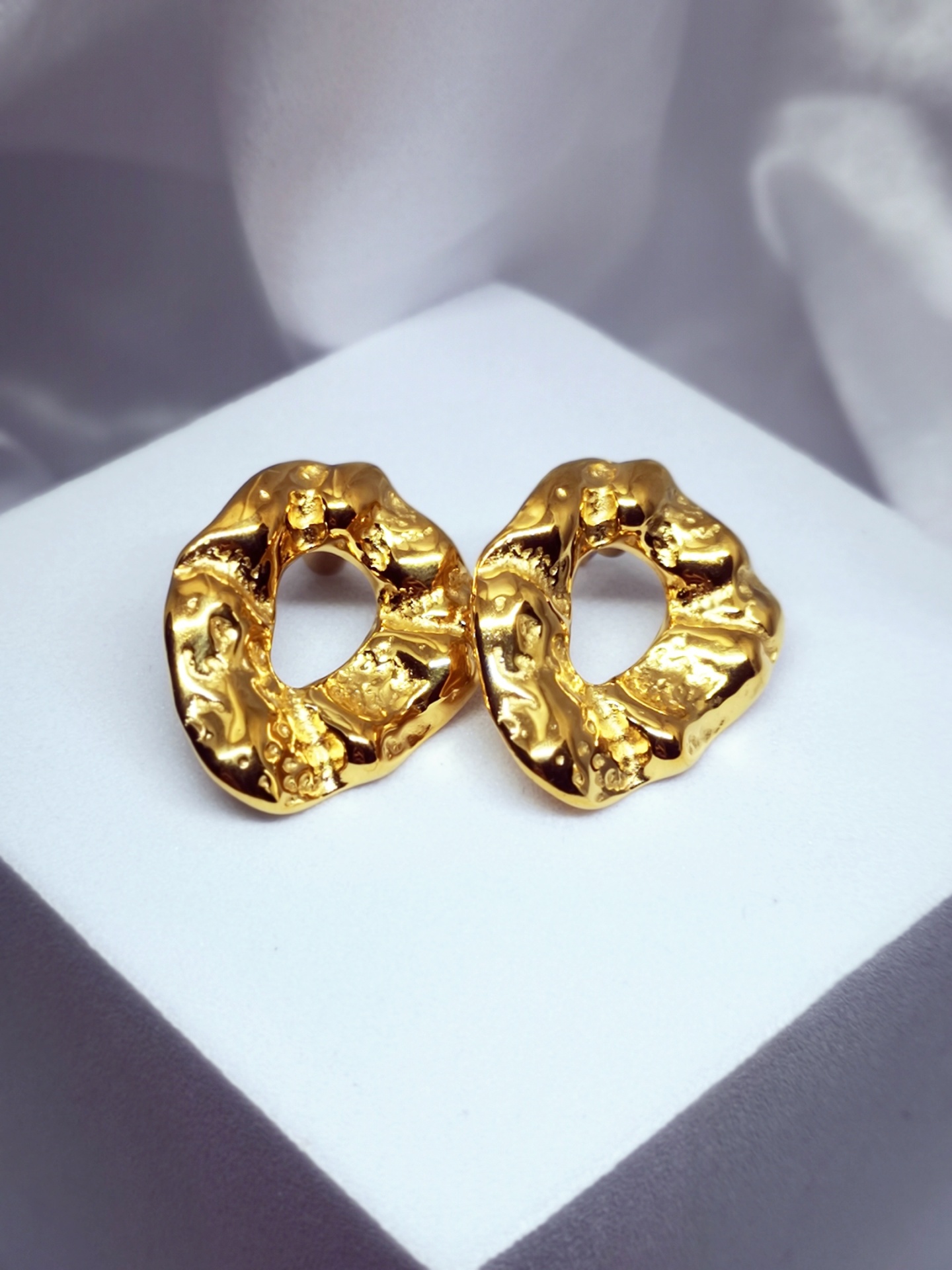 Brincos dourados texturizados em forma irregular com abertura central em fundo branco e cinza