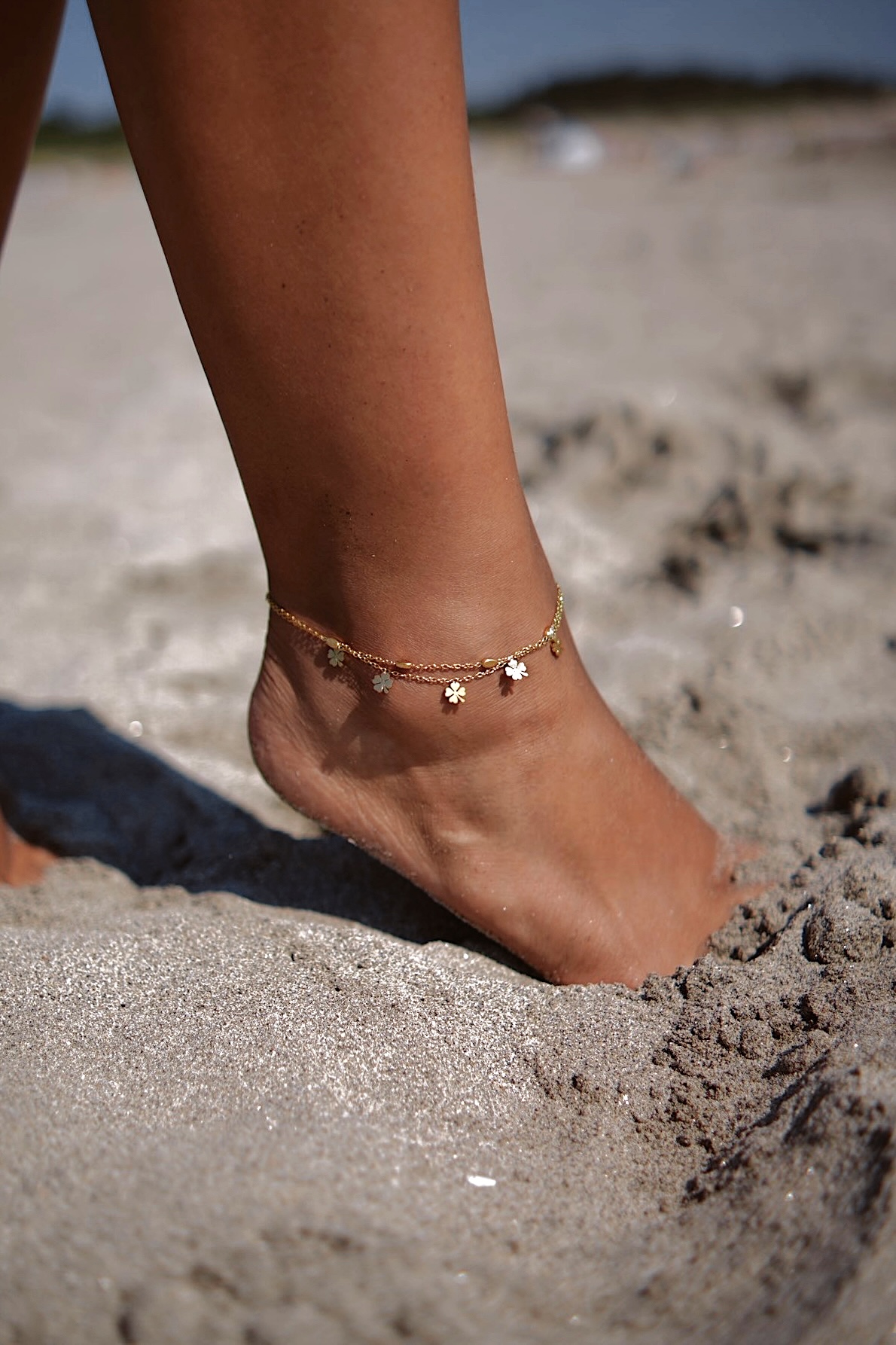 Tornozeleira dourada com pendentes de flores numa perna na areia da praia