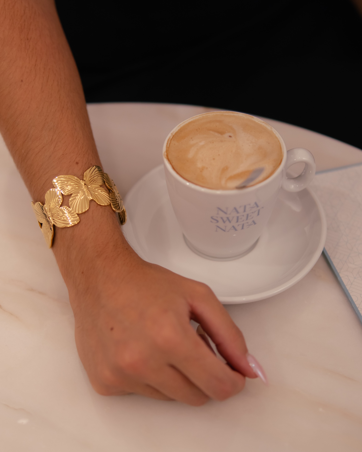 Pulseira dourada de folhas em metal e chávena branca de café com leite numa mesa de mármore