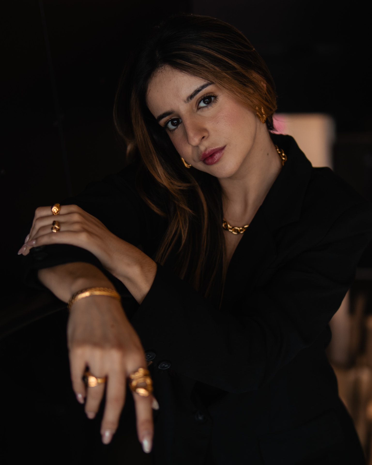 Mulher com joias douradas e blazer preto, em fundo escuro.