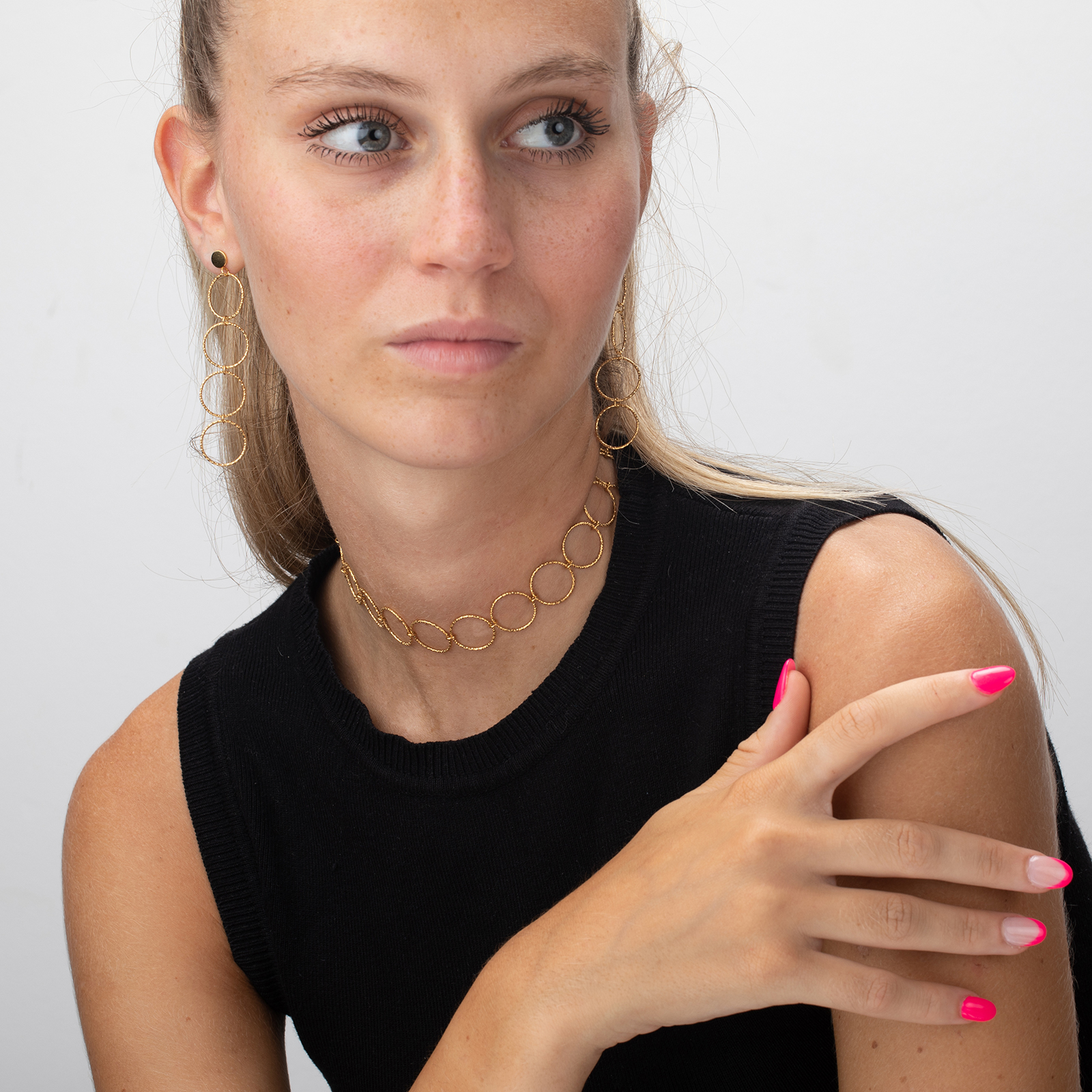 Mulher com brincos e gargantilha dourados, camisa preta sem mangas e unhas rosa.