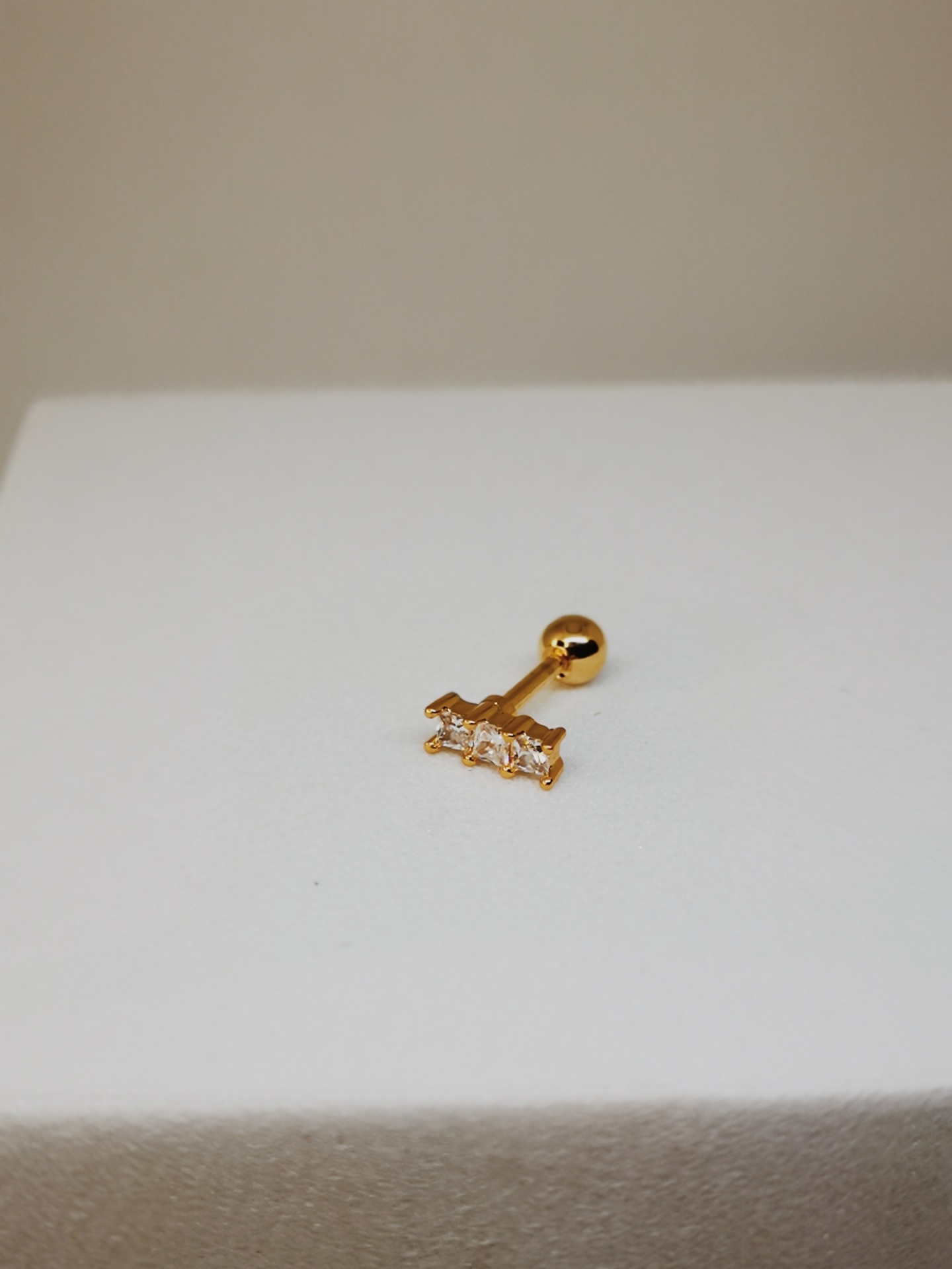 Piercing dourado com três pedras retangulares sobre superfície branca