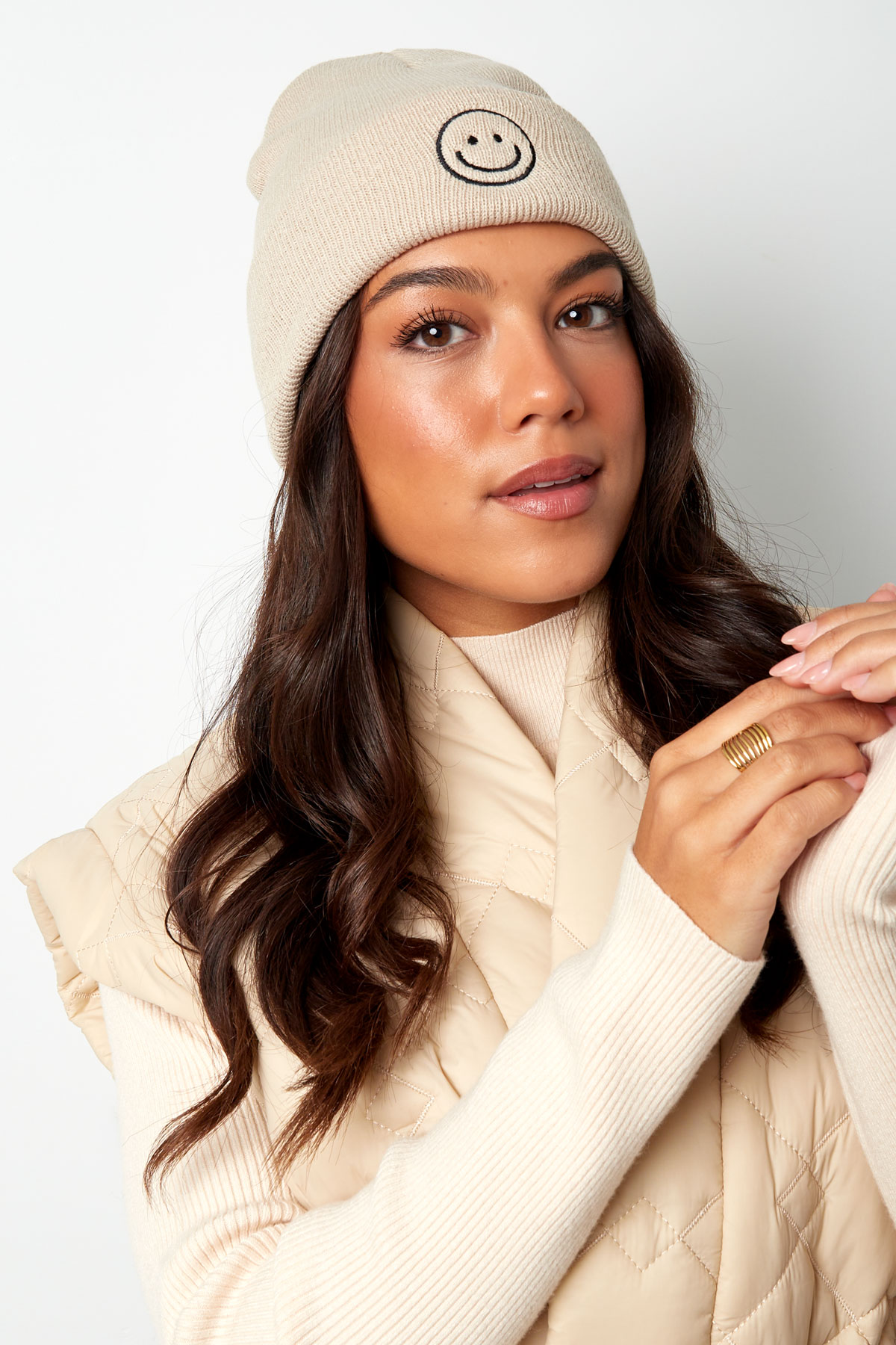 Gorro de malha bege com rosto sorridente bordado, usado por mulher com casaco e camisola bege.