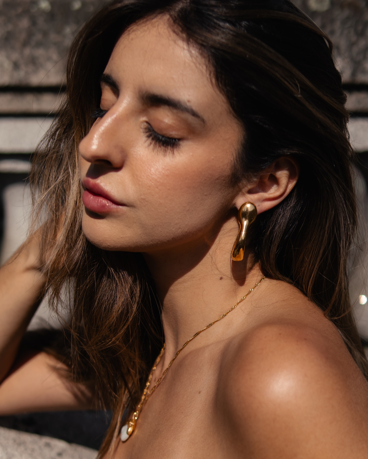 Mulher com brincos dourados e colar em fundo rochoso desfocado
