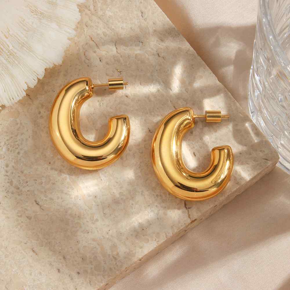 Brincos dourados em forma de arco sobre pedra cremosa com copo transparente ao lado