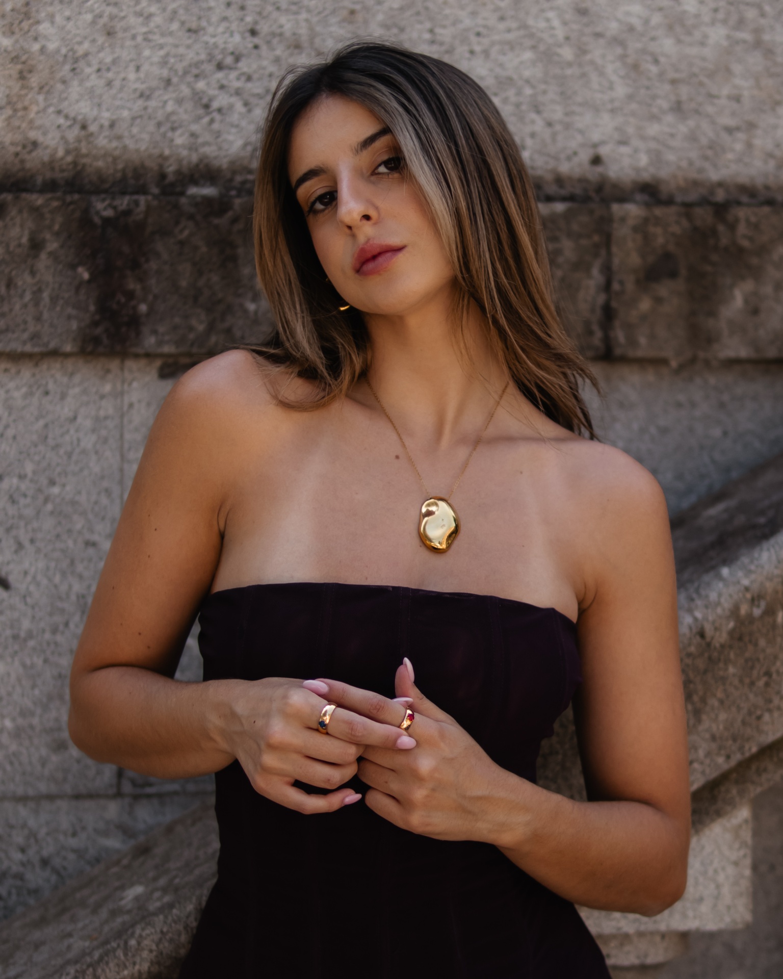 Mulher com colar dourado e anéis, vestido preto, em fundo de muro de pedra.