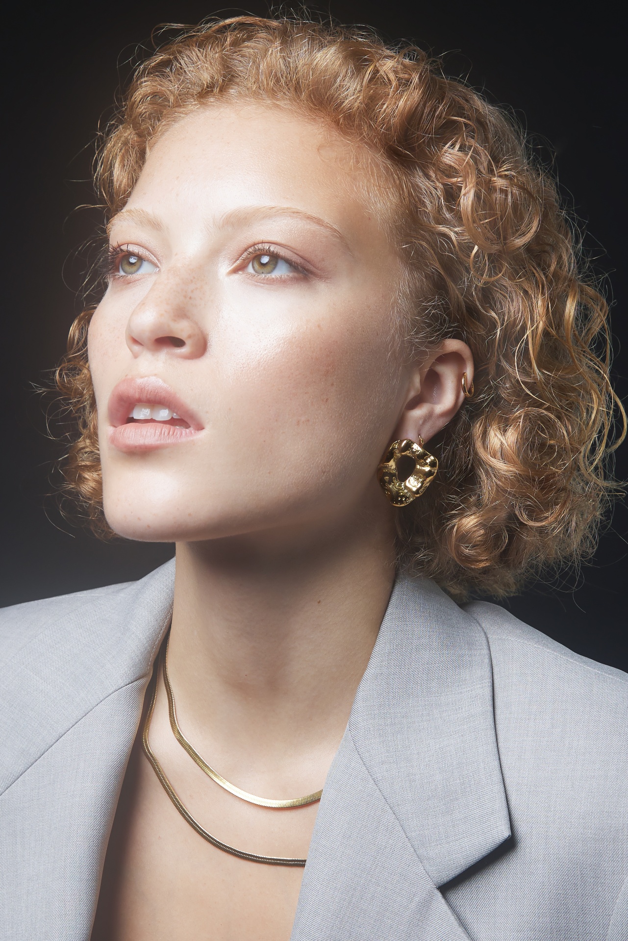 Mulher com brincos e colares dourados e blazer cinza claro