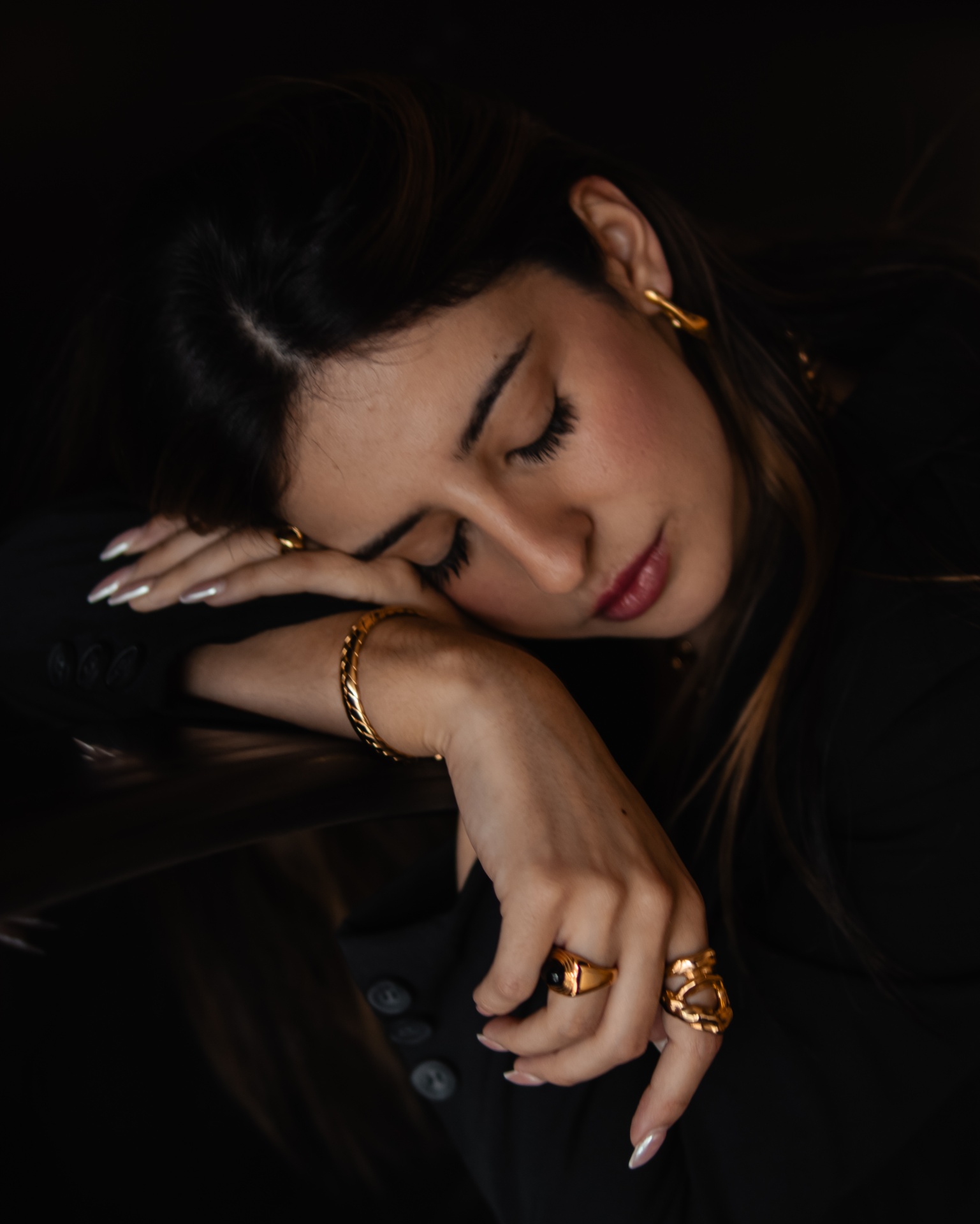 Mulher com acessórios dourados e casaco preto sobre fundo escuro