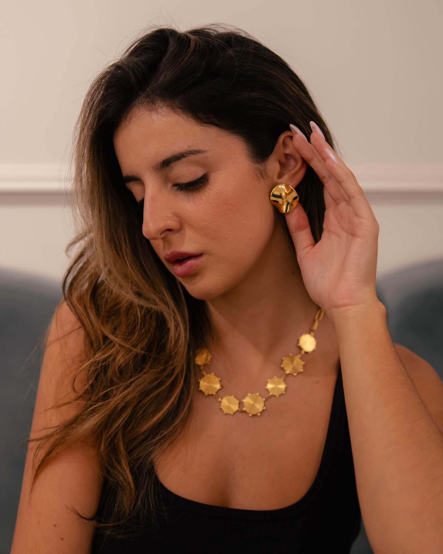 Mulher com colar e brinco dourados, vestindo top preto, em ambiente interior.