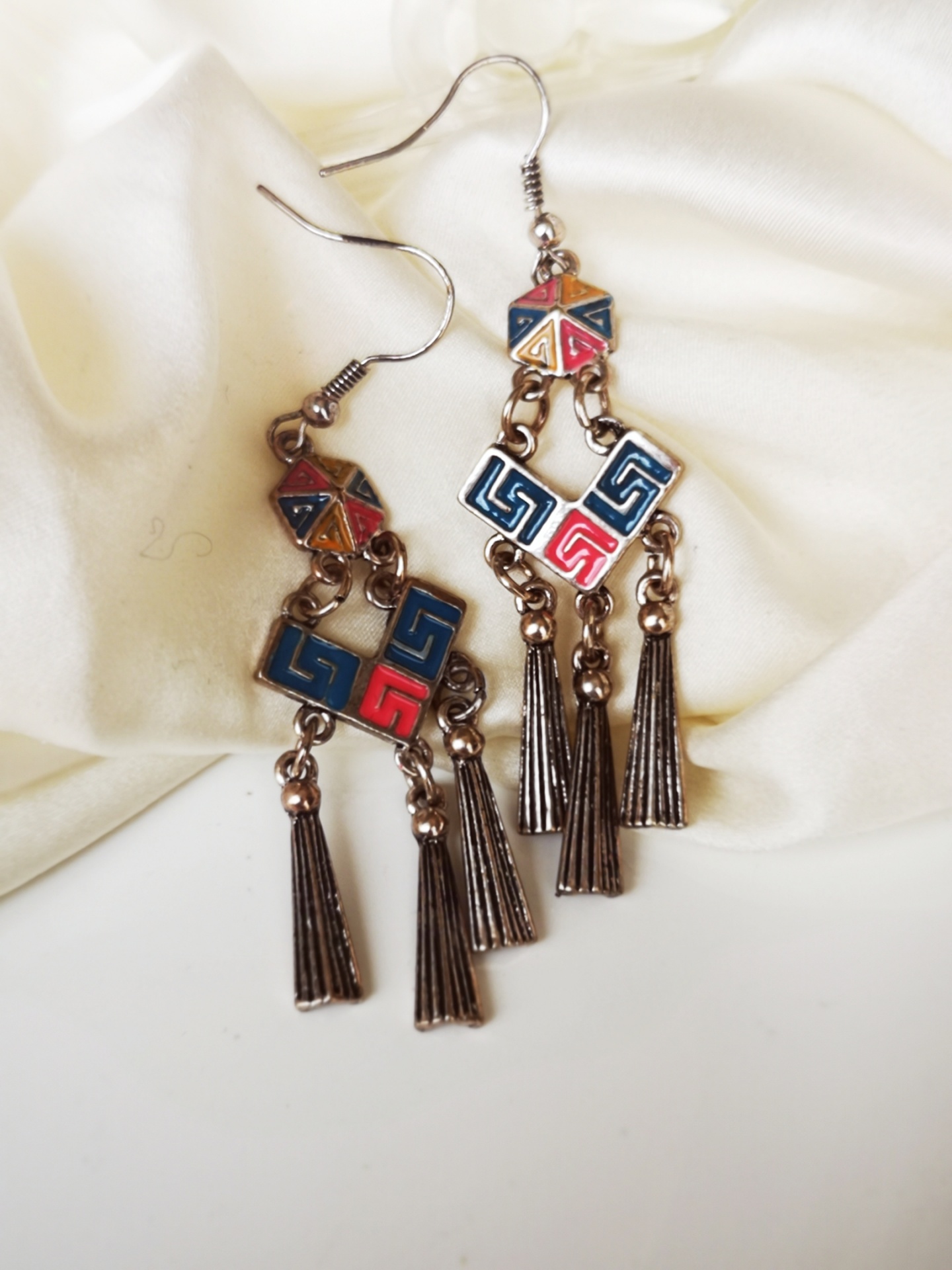 Brincos pendentes com padrões geométricos coloridos e pendentes metálicos sobre tecido branco