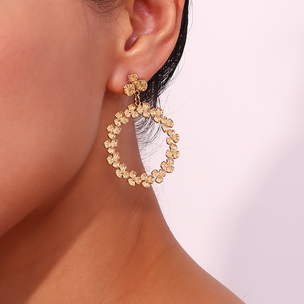 Brinco dourado em argola com flores pequenas, usado na orelha de uma pessoa, fundo rosa claro