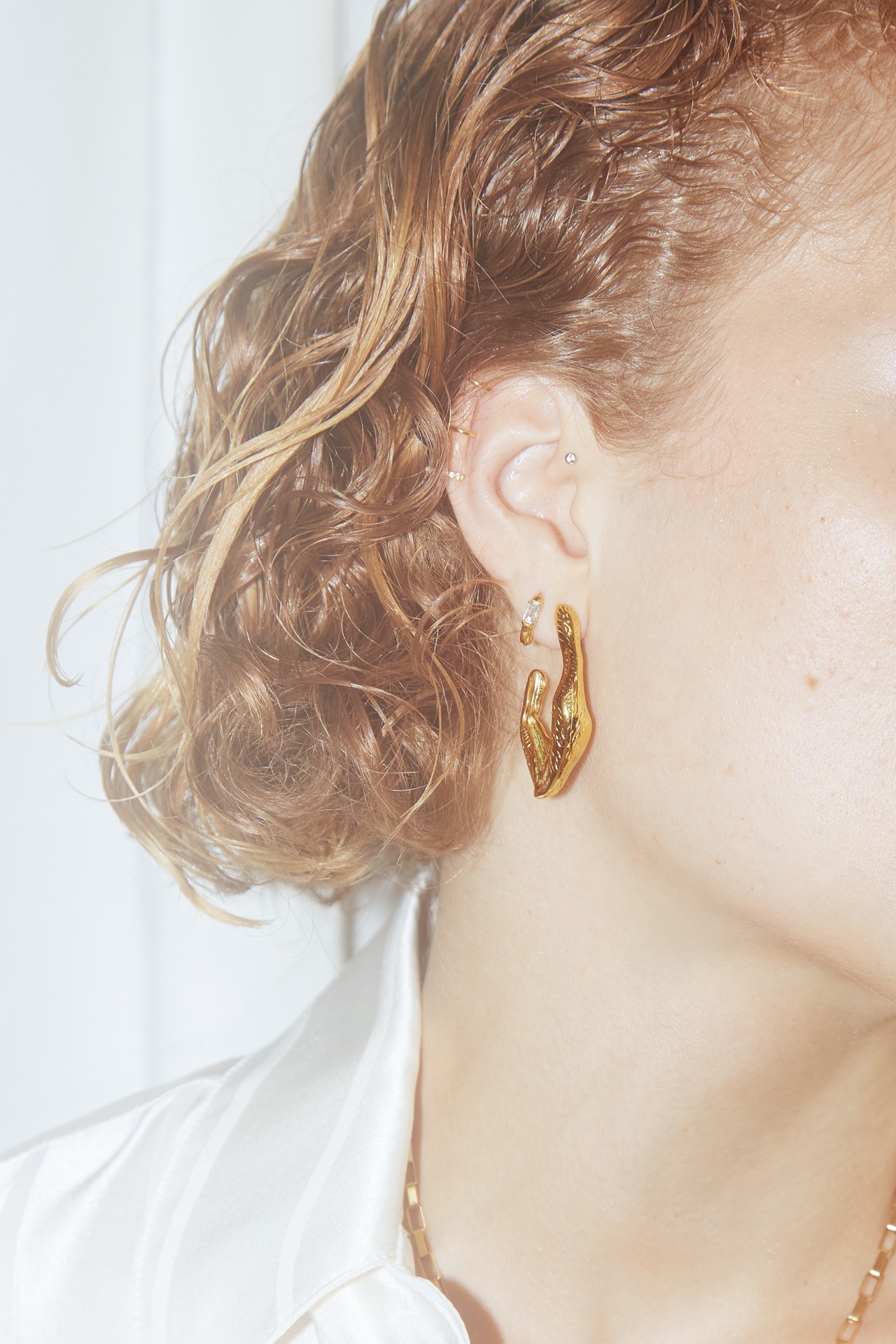 Brincos dourados com formato orgânico em orelha de pessoa com cabelo castanho claro ondulado e camisa branca.