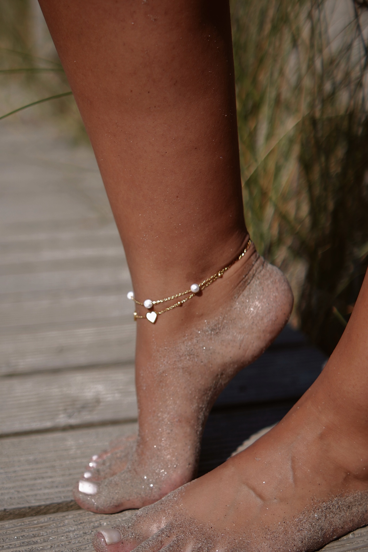 Tornozelo feminino com bracelete dourada dupla com pérolas e coração, pés com areia, passadiço de madeira