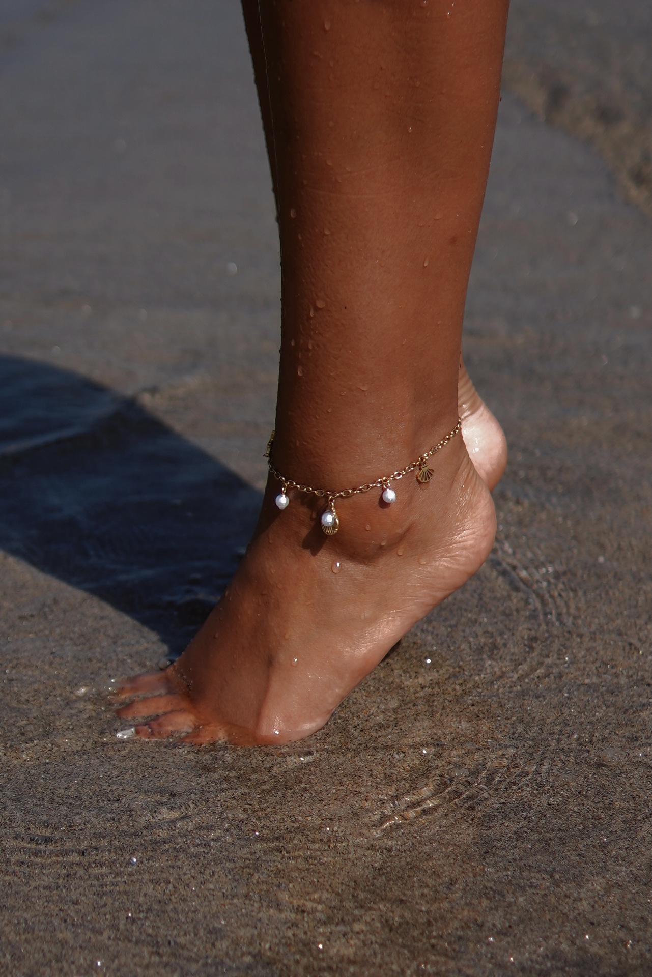 Pé com tornozeleira dourada com pendentes de pérolas na areia molhada da praia