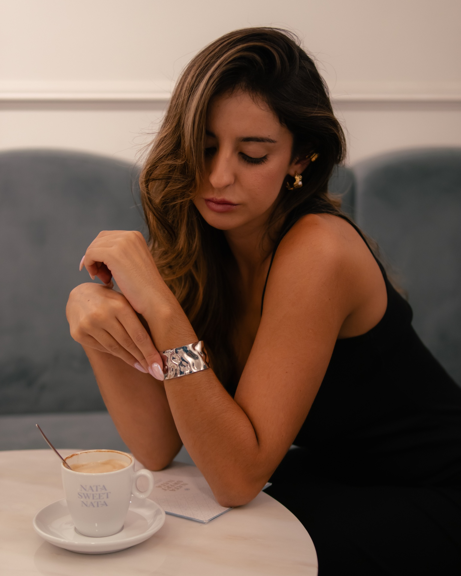 Mulher com vestido preto e pulseira prateada sentada à mesa com chávena branca de café