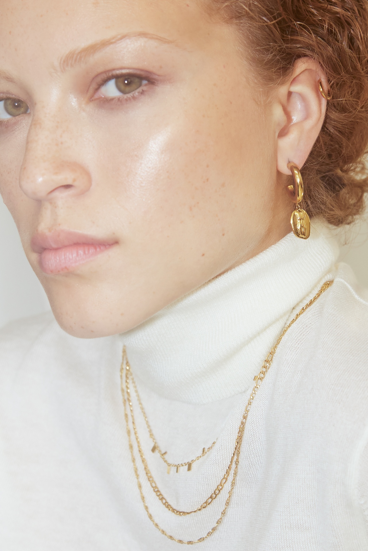 Mulher com brincos e colares dourados, camisola branca de gola alta