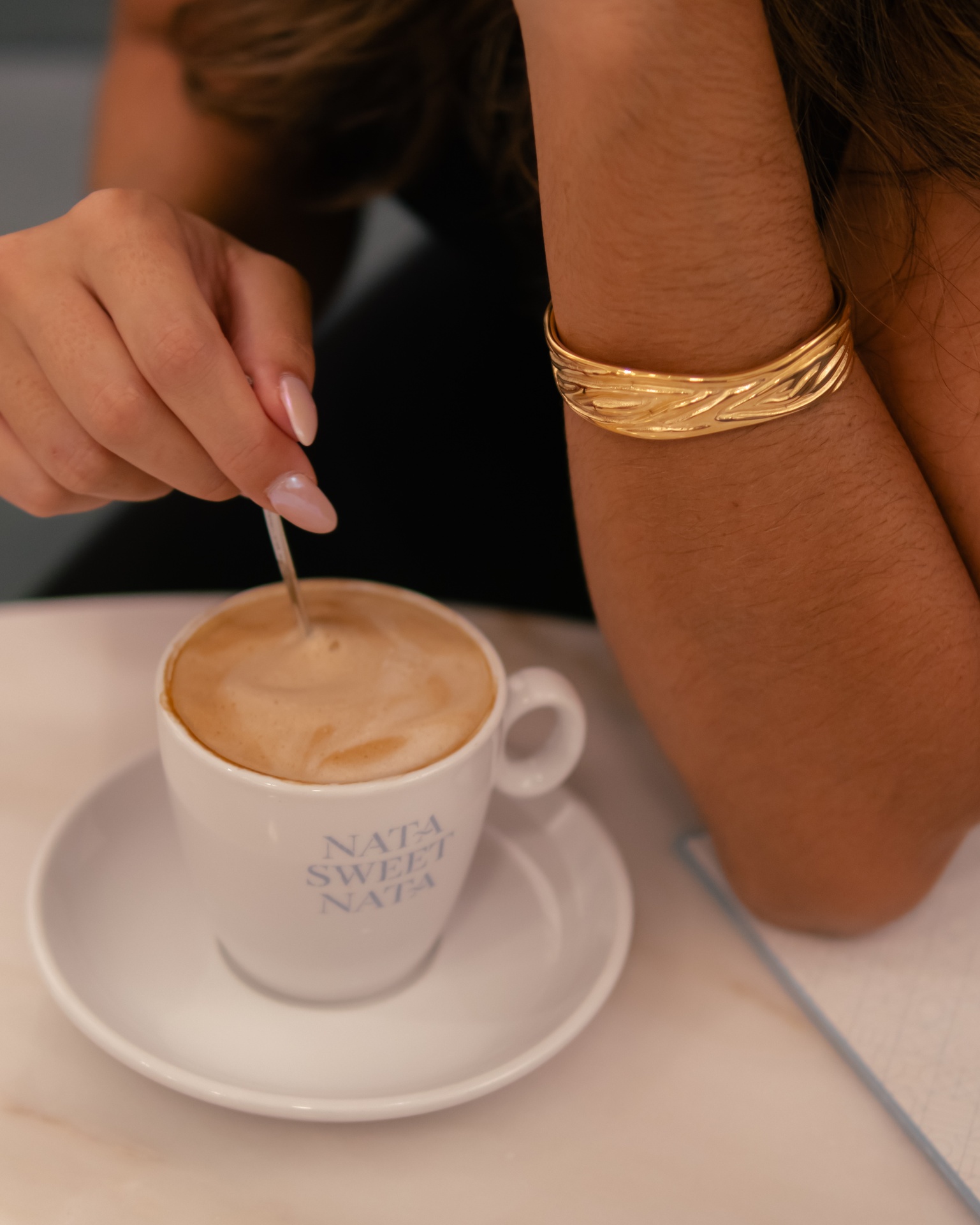 Taça branca de café com texto, mão feminina mexendo com colher e pulseira dourada no pulso