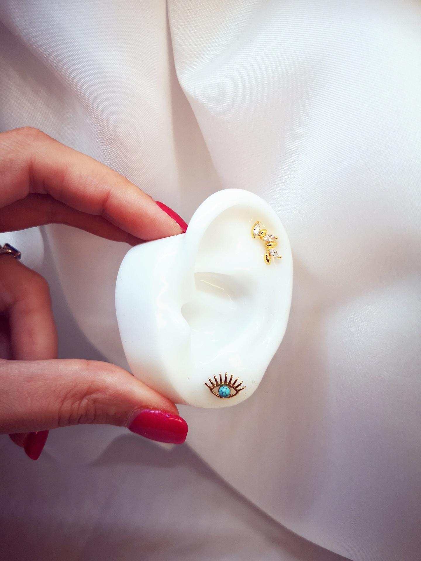 Peça em forma de orelha branca com dois brincos dourados, segurada por mão com unhas vermelhas.