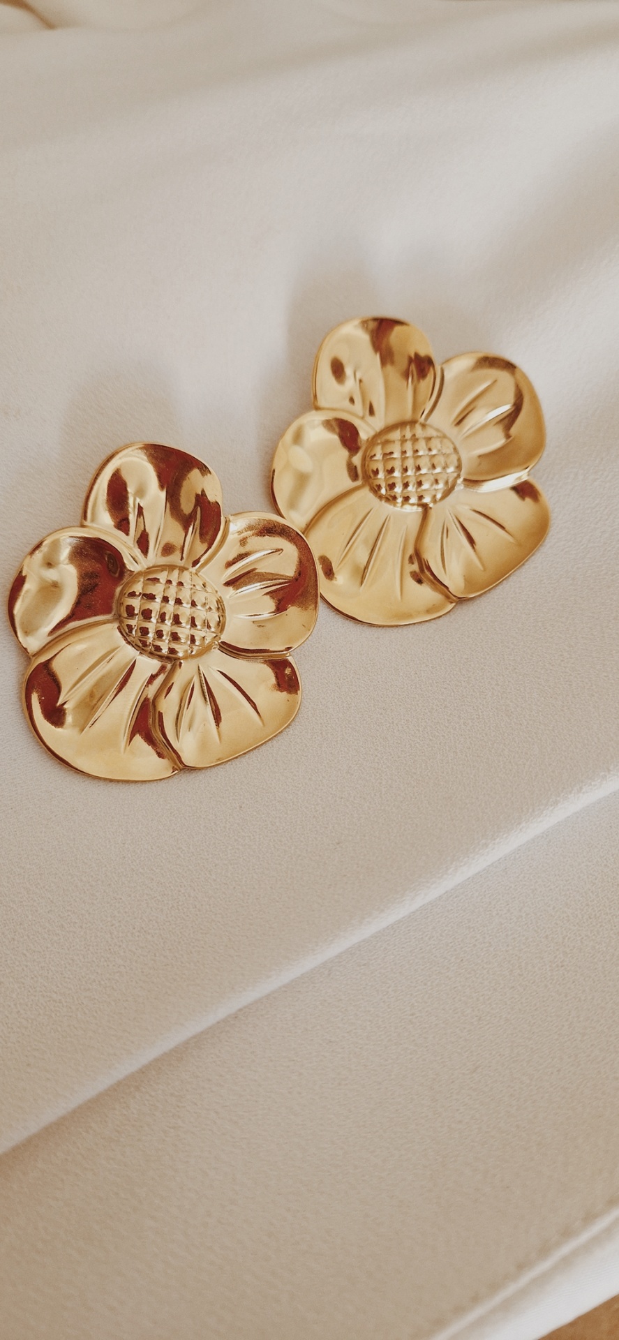 Brincos dourados em forma de flores com textura no centro, sobre tecido branco.