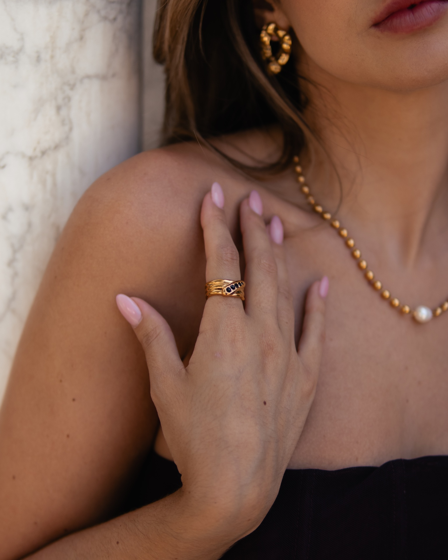 Anel dourado com pedra escura na mão de mulher com acessórios dourados e fundo de parede clara
