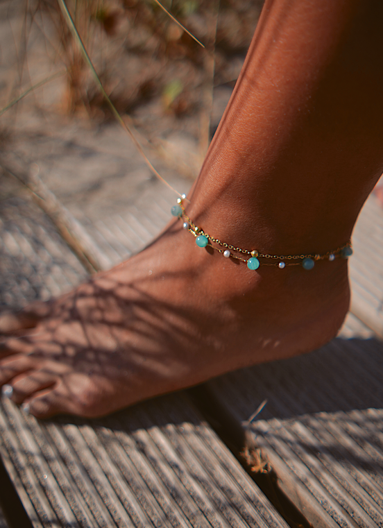 Tornozeleira dourada com contas azuis e brancas num tornozelo sobre madeira clara